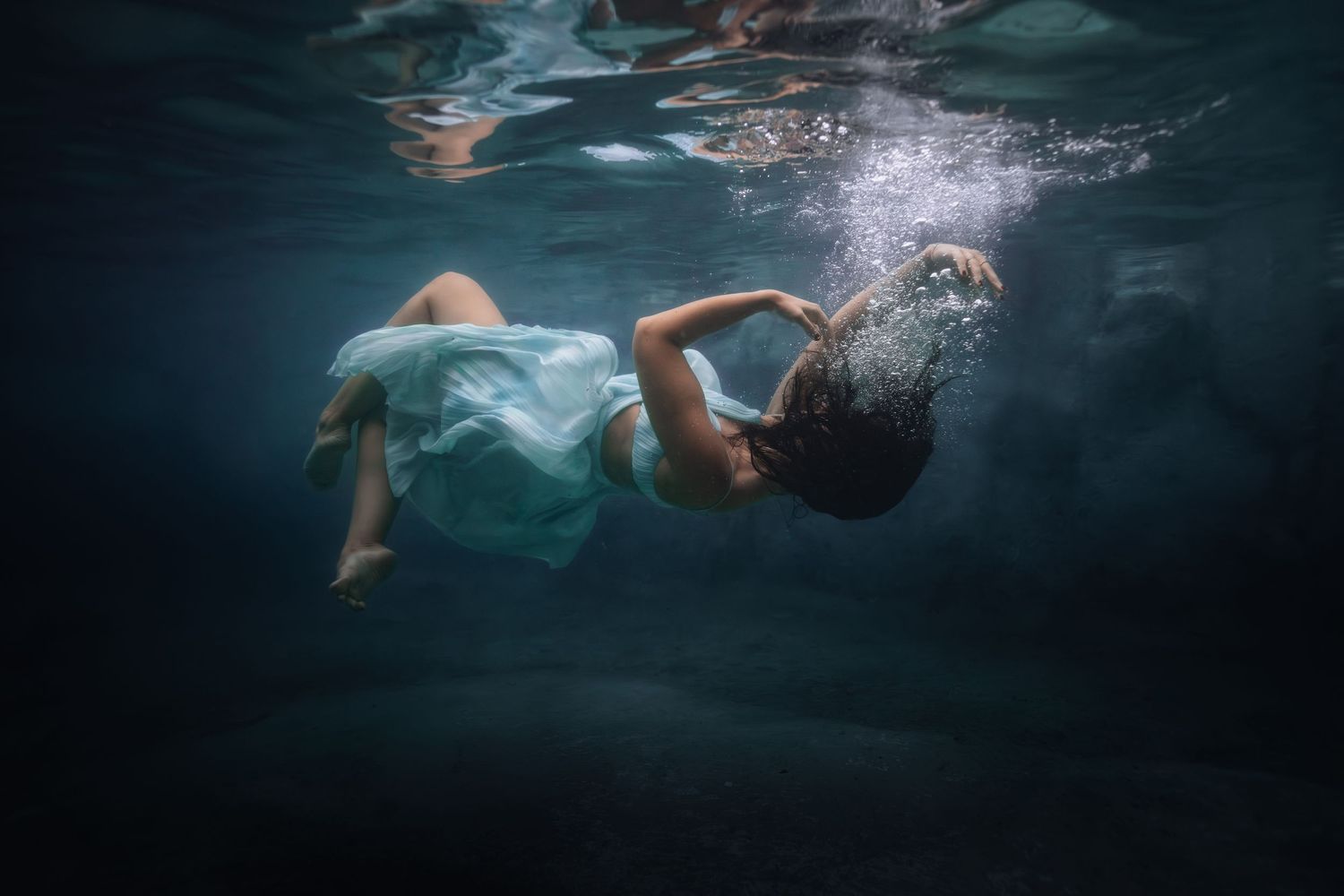 Underwater view of a graceful swimmer in a teal dress gliding through dark blue water creating ethereal ripples.