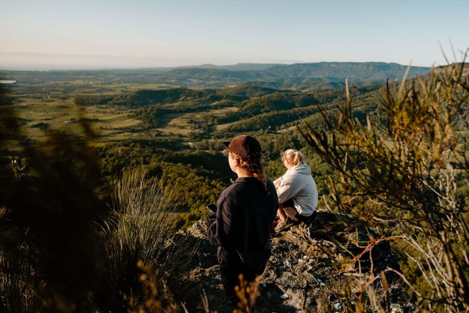 Two people sit on a rocky mountain lookout enjoying panoramic views of lush green valleys below at golden hour.