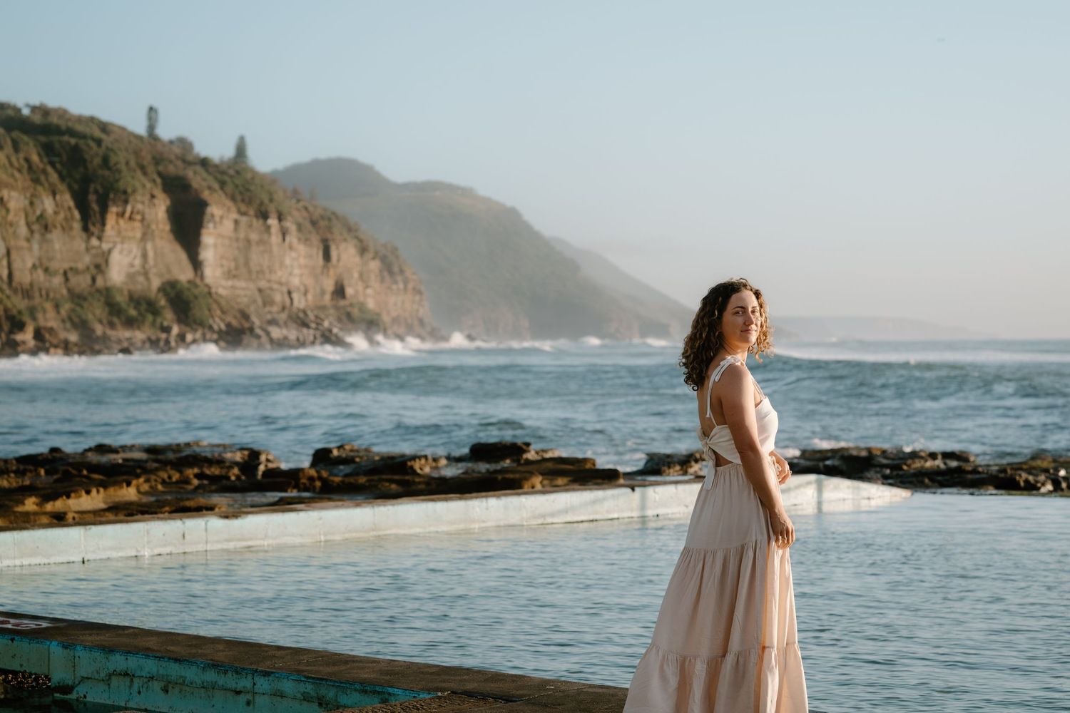 Woman in flowing pink dress poses by coastal rock pool with dramatic cliffs and ocean waves in background.