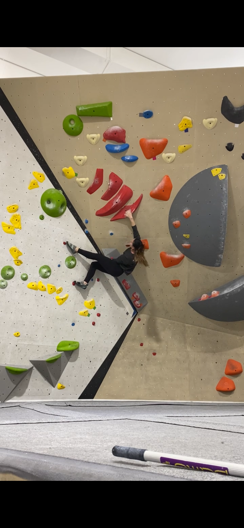 A climber scales a colorful indoor bouldering wall with various holds in an indoor climbing gym.