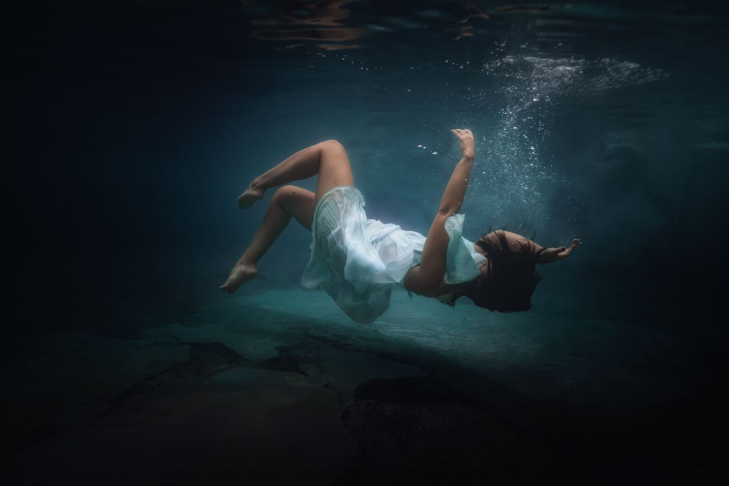A person swimming underwater in a dark blue pool with dramatic lighting creating a moody underwater scene.