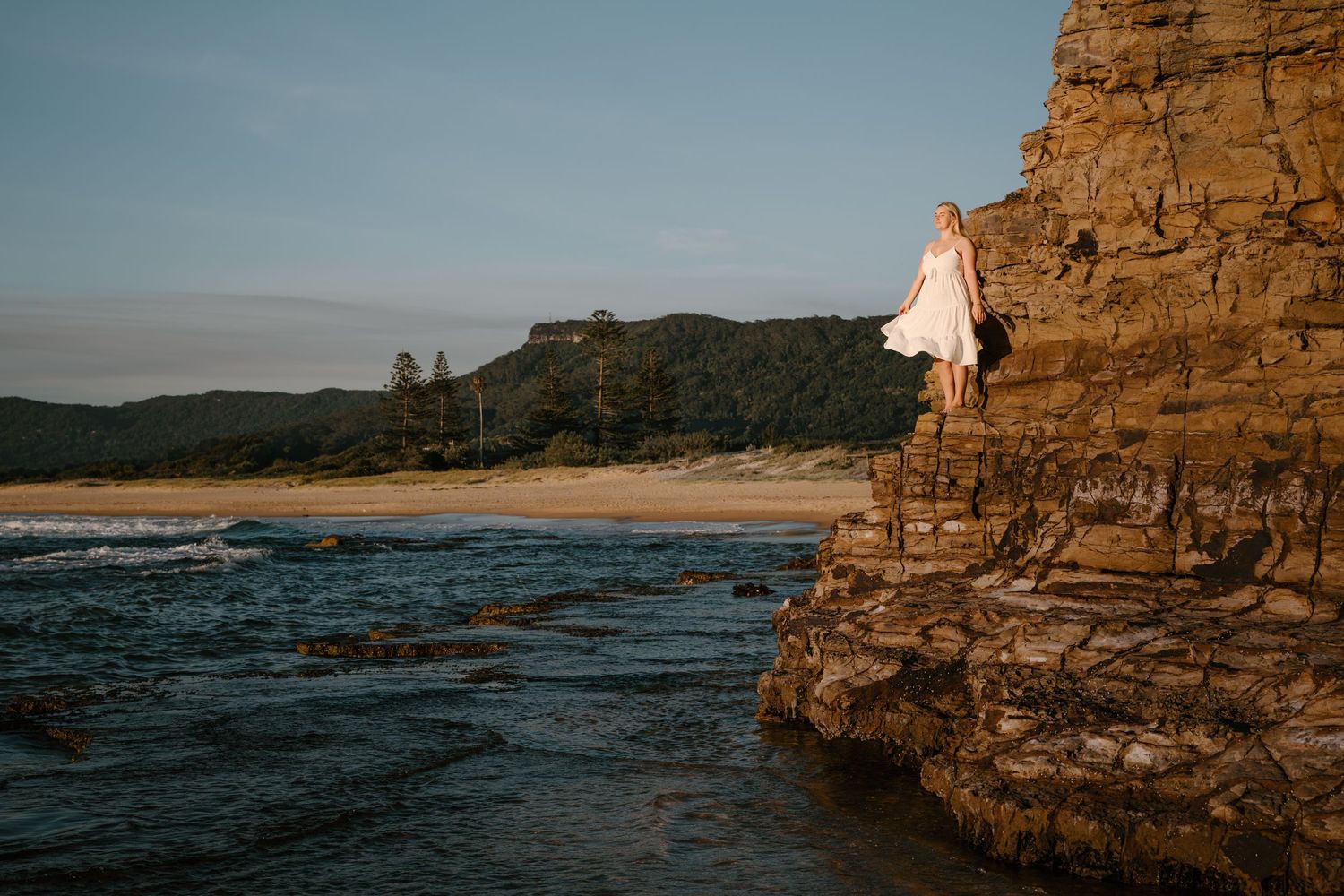 Scenic coastal landscape with someone sitting on rocky cliff edge overlooking beach and ocean at sunset.