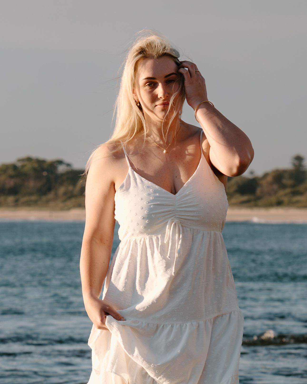A person in a flowing white sundress poses on a beach at sunset with ocean waves and coastline in the background.
