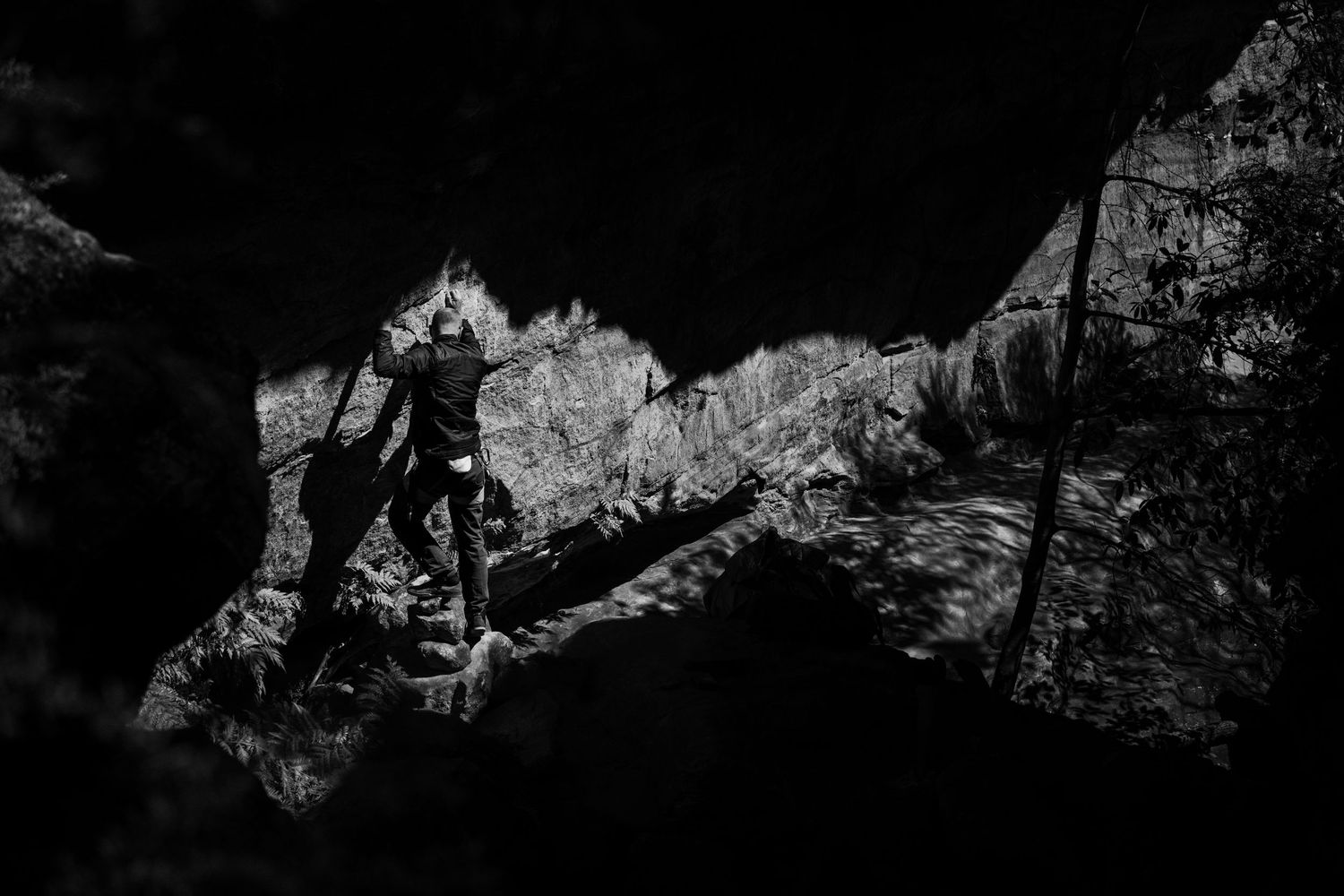 Black and white photo of a climber silhouetted in a dark cave opening with rocky mountain landscape behind.