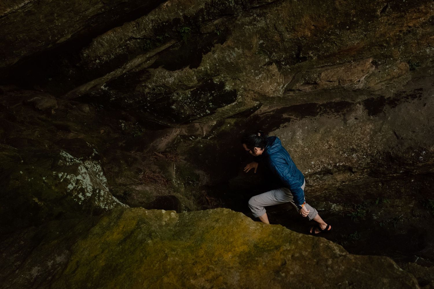 Person in blue jacket rock climbing on dark stone boulders at night with headlamp illuminating the surface.
