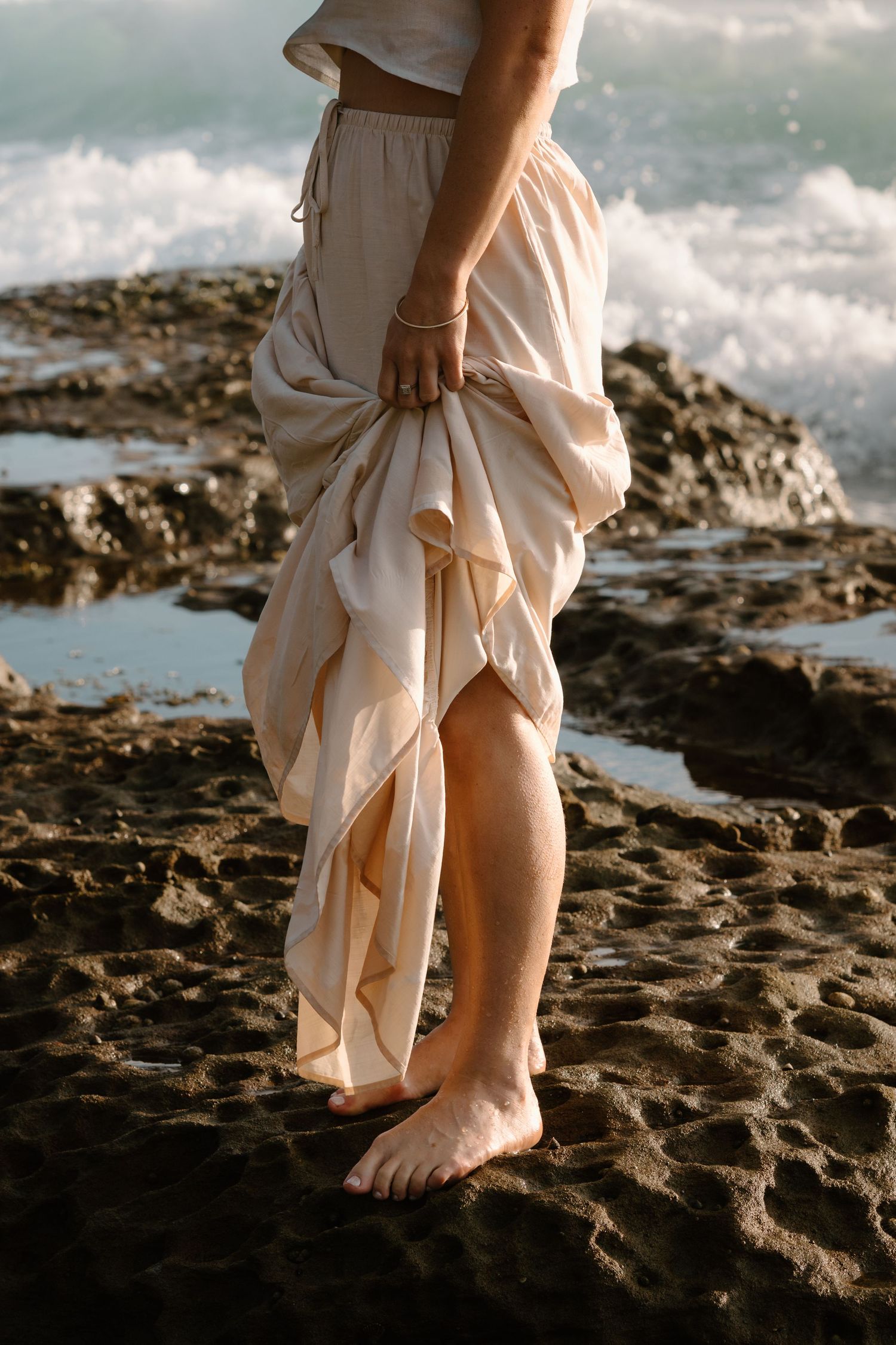 Bare feet walking on rocky coastal shore in flowing white dress against dramatic ocean waves and cloudy sky.