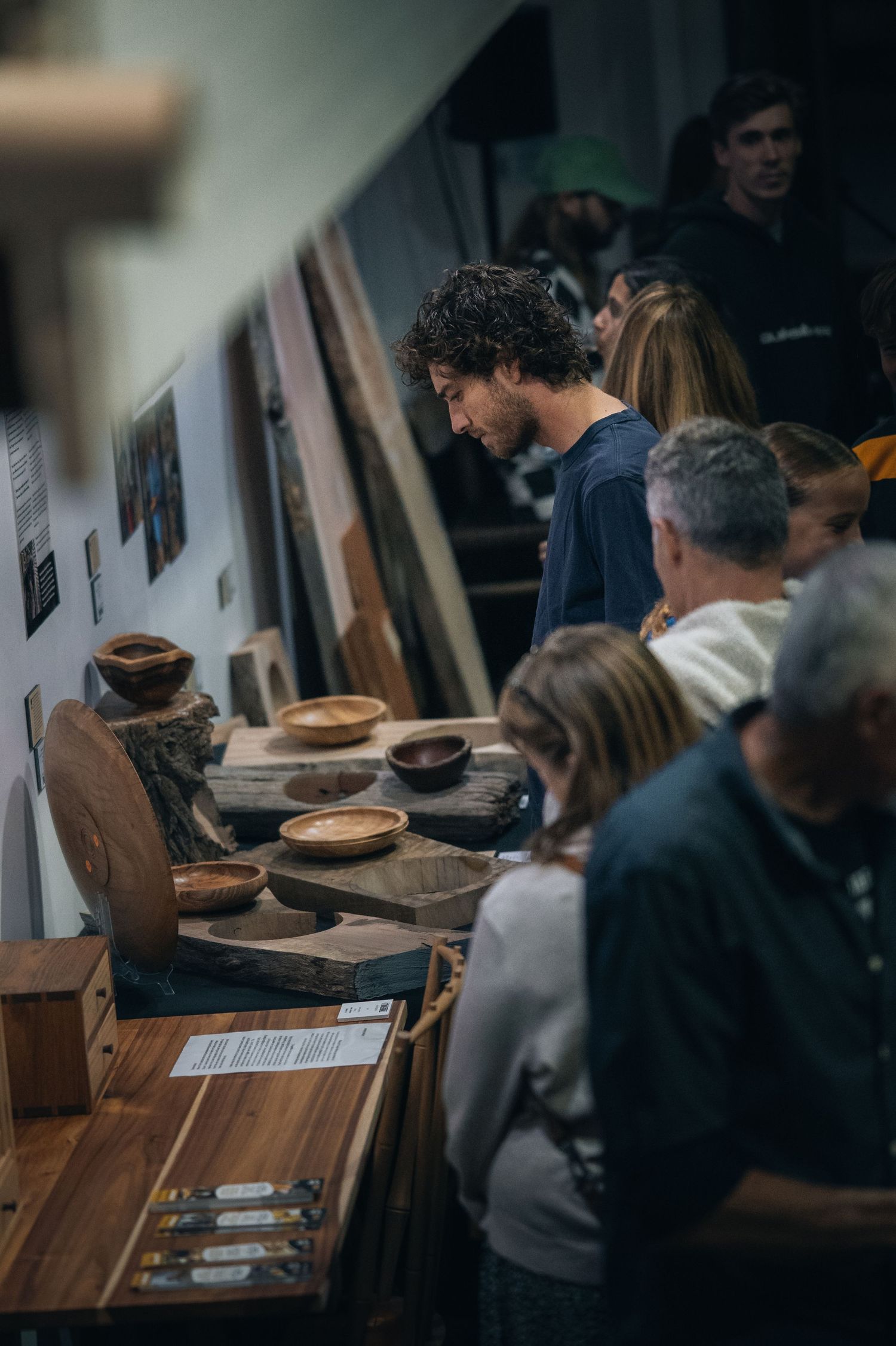 People browse wooden bowls and handcrafted items on display at a rustic artisan market or gallery exhibition.