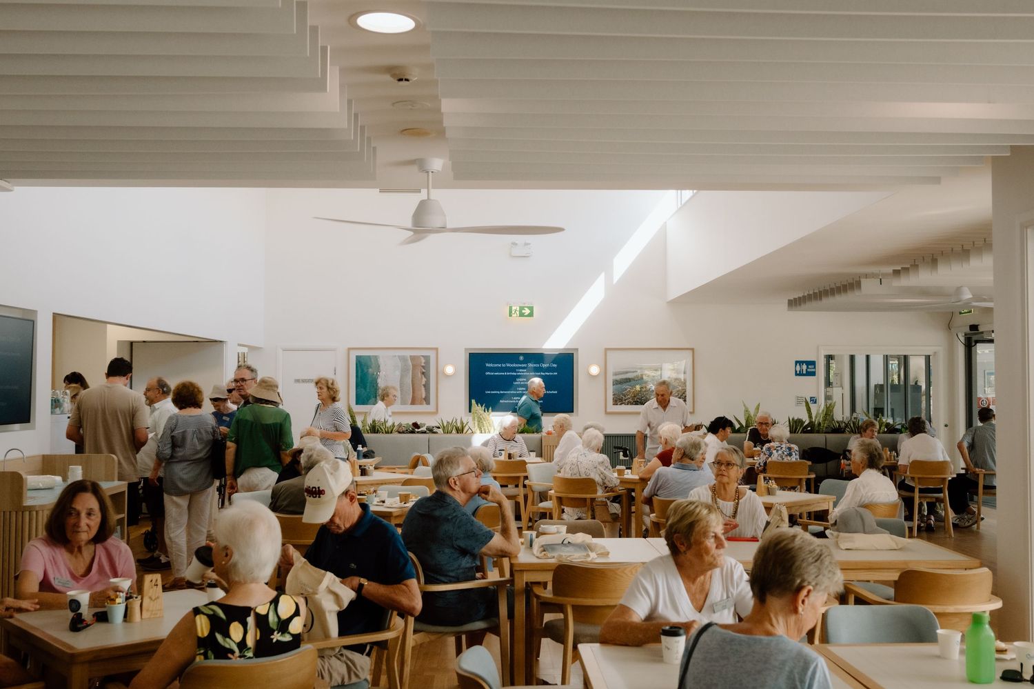 Busy café dining room with patrons seated at wooden tables enjoying meals in a bright, white-walled restaurant.