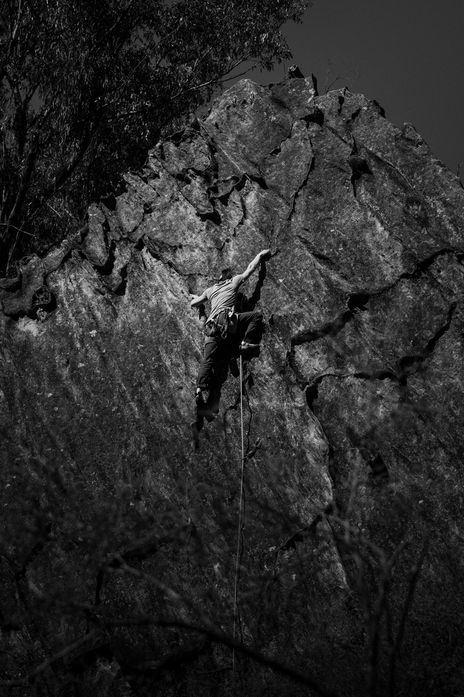 Black and white photo of a rock climber scaling a rugged cliff face with a rope in dramatic lighting.