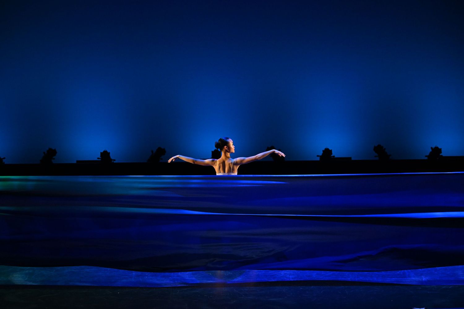 Swimmer emerges from glowing blue pool at night, arms spread wide against a dramatic dark blue backdrop.