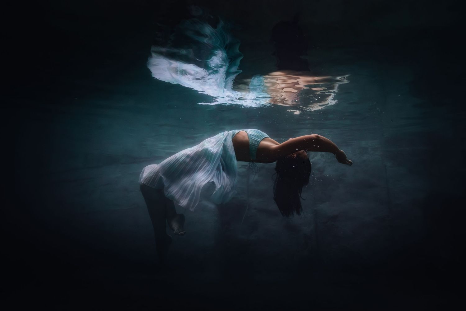 Ethereal underwater photograph showing a figure in flowing white fabric gracefully floating in dark turquoise waters.