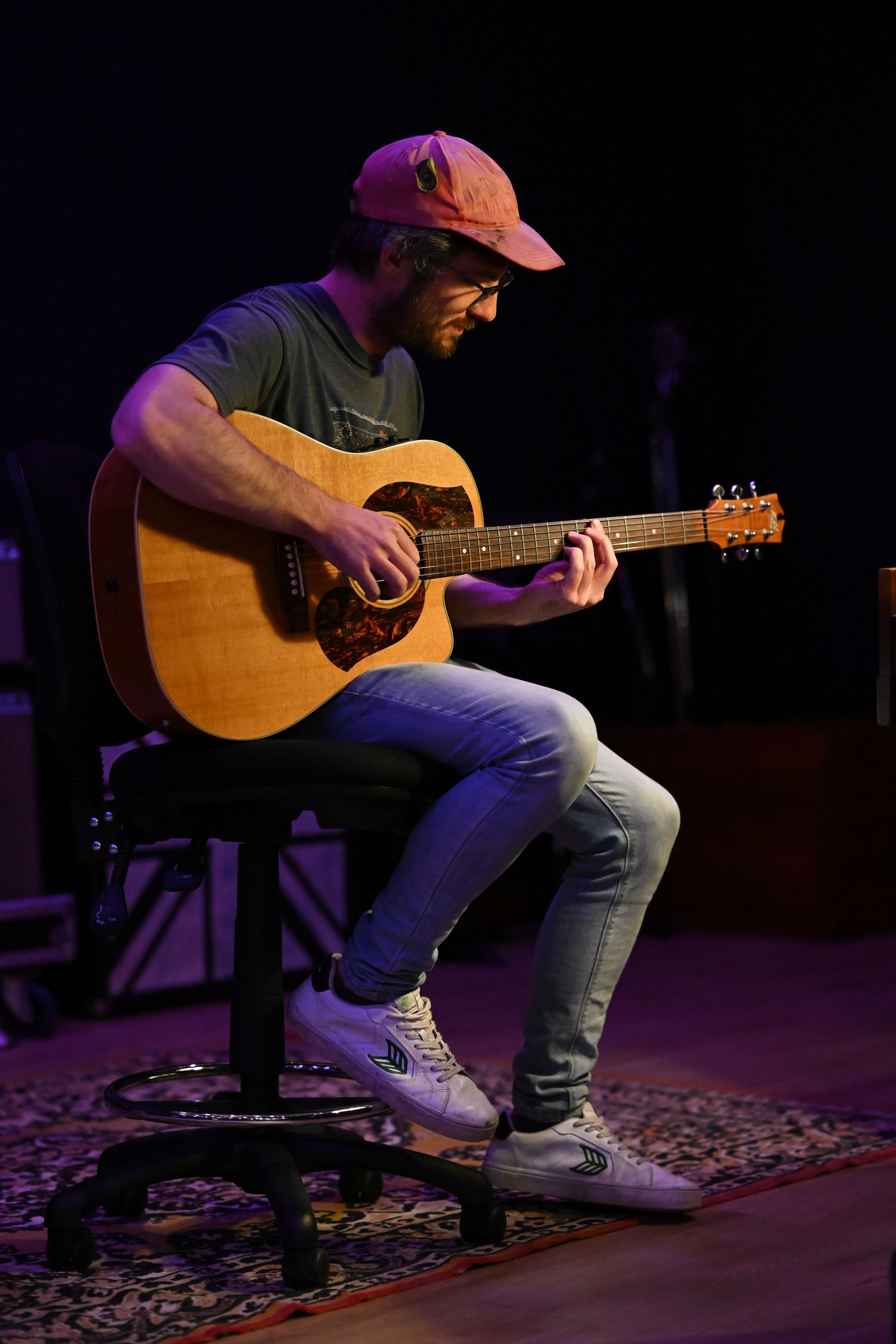 A musician performs on an acoustic guitar while seated on a stool under purple stage lighting.