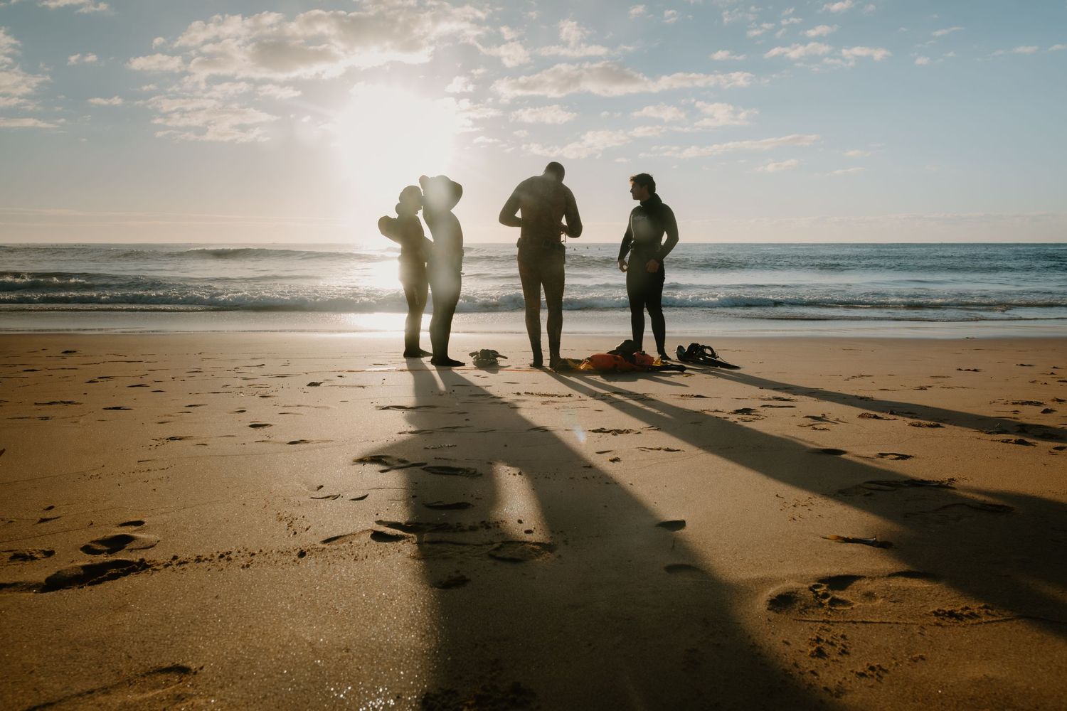 Three people stand on a sandy beach at sunset, casting long shadows with ocean waves in the background.