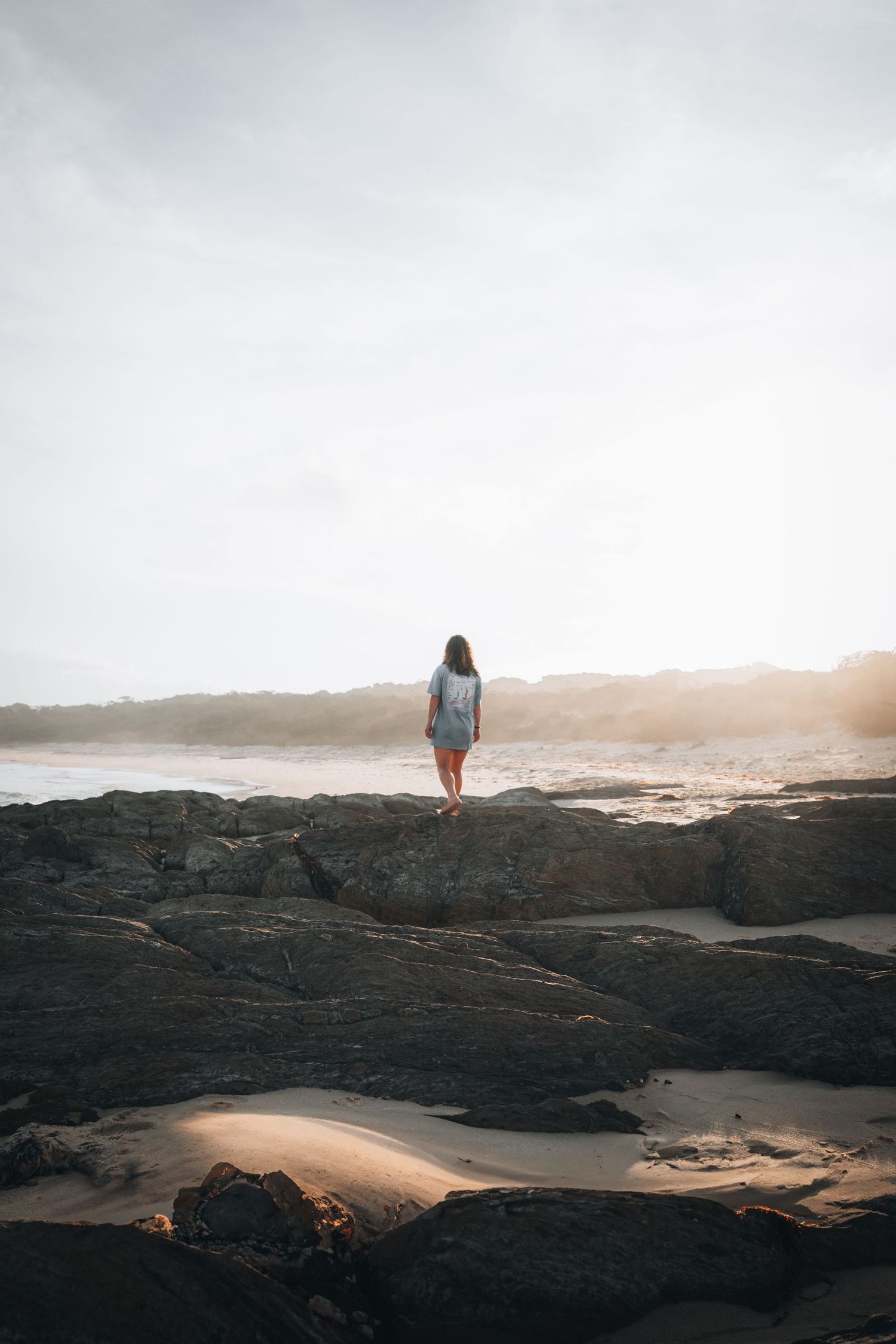 A solitary figure stands on dark volcanic rocks overlooking a misty beach shore at sunset.