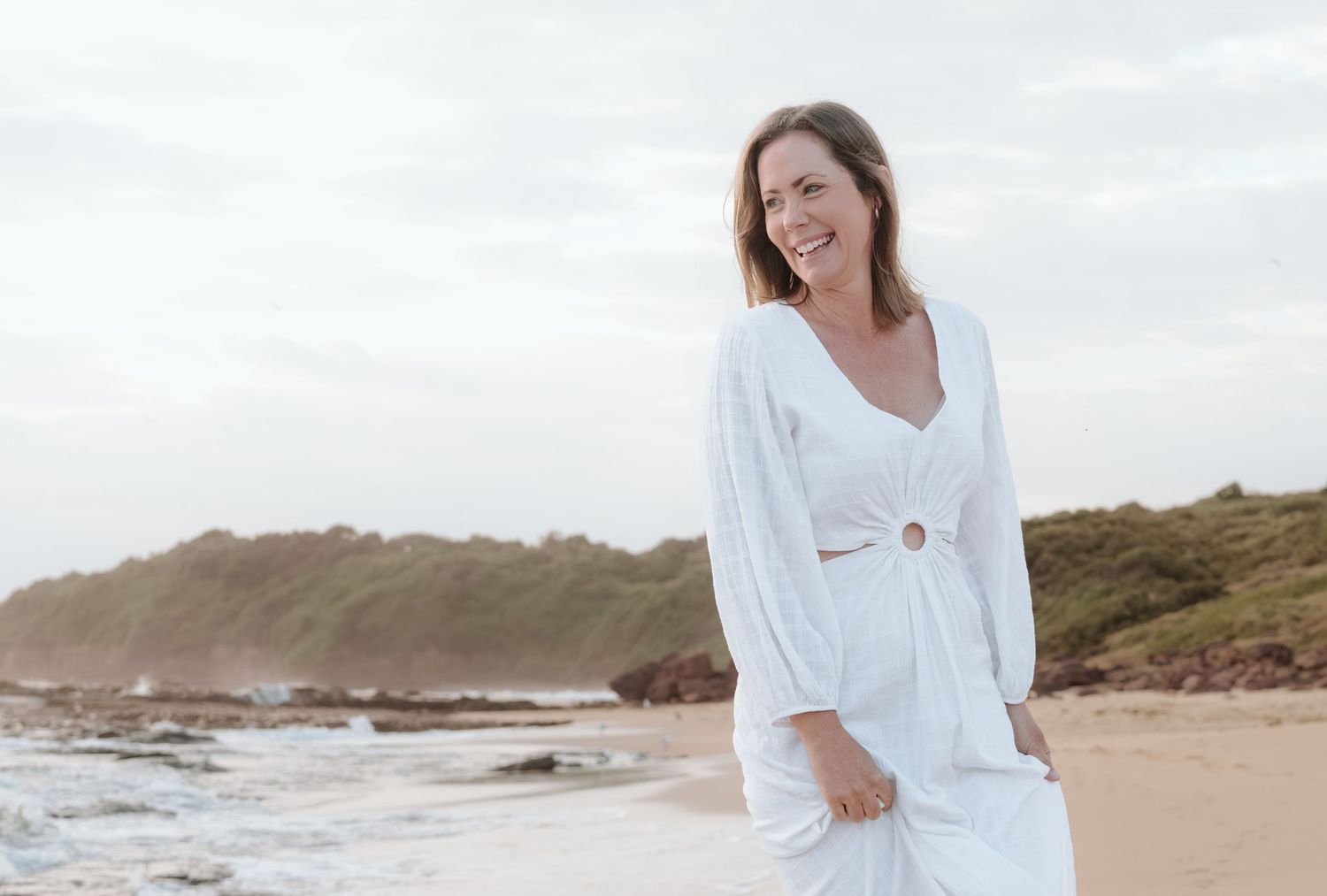 Person in flowing white dress stands on sandy beach with coastal cliffs in background under cloudy sky.