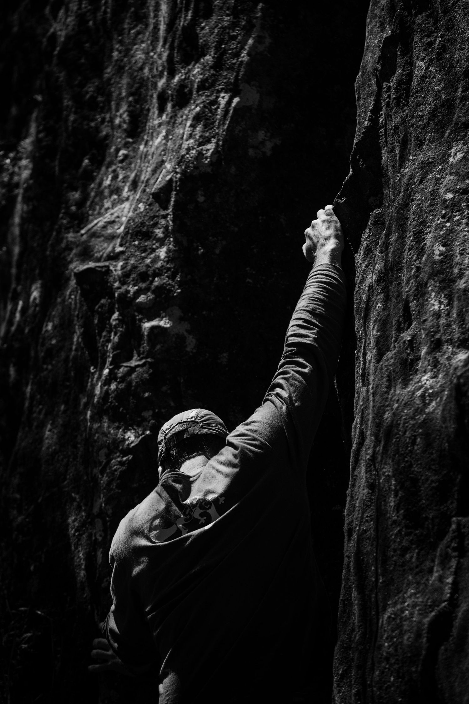 Black and white photo of a rock climber gripping a narrow cliff face crevice, reaching upward with determination.