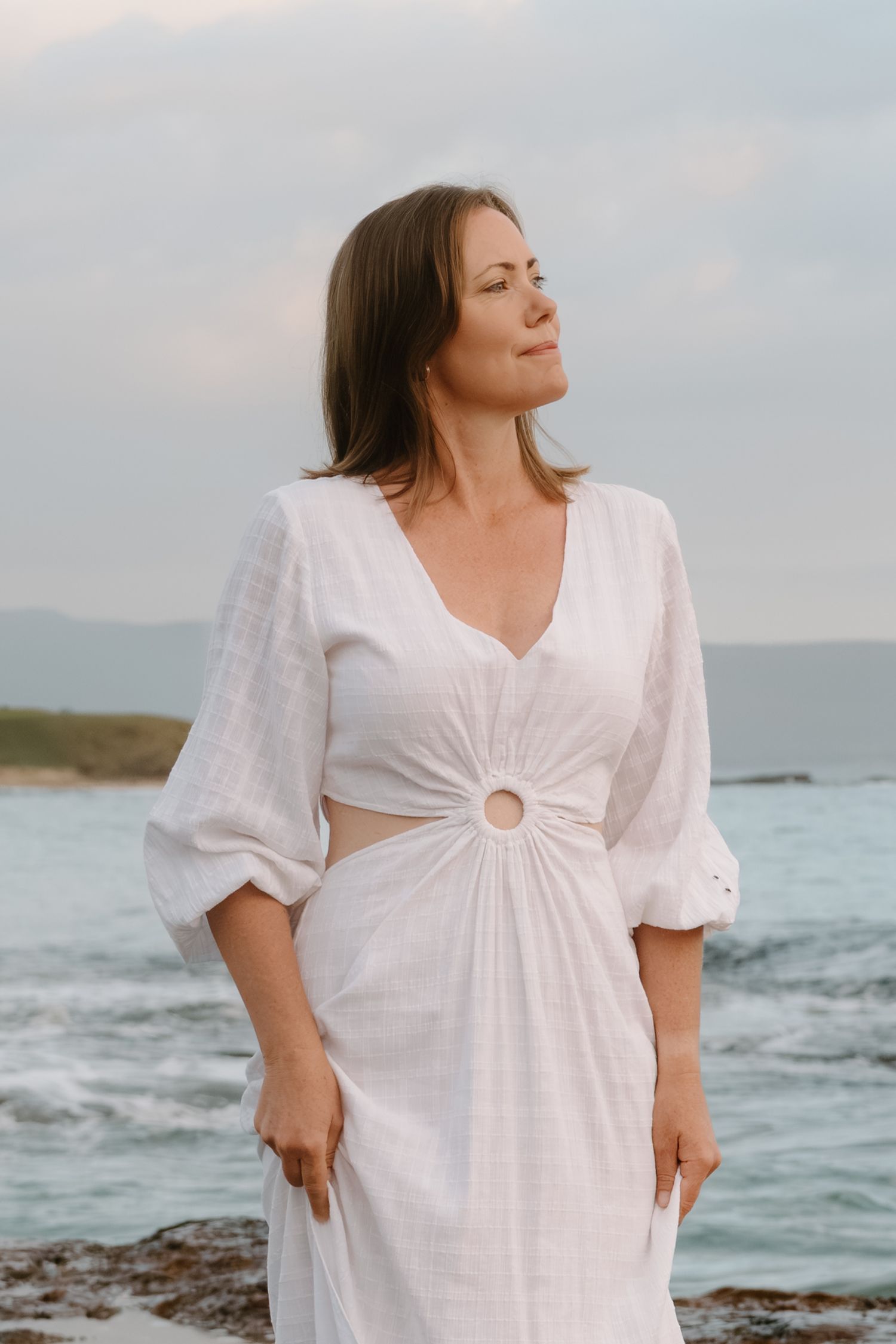 Woman in flowing white dress poses gracefully against ocean backdrop during sunset photoshoot at beach.