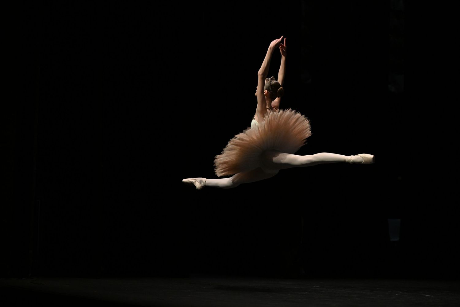 A ballerina in a beige tutu performs a graceful grand jeté leap against a dark stage background.
