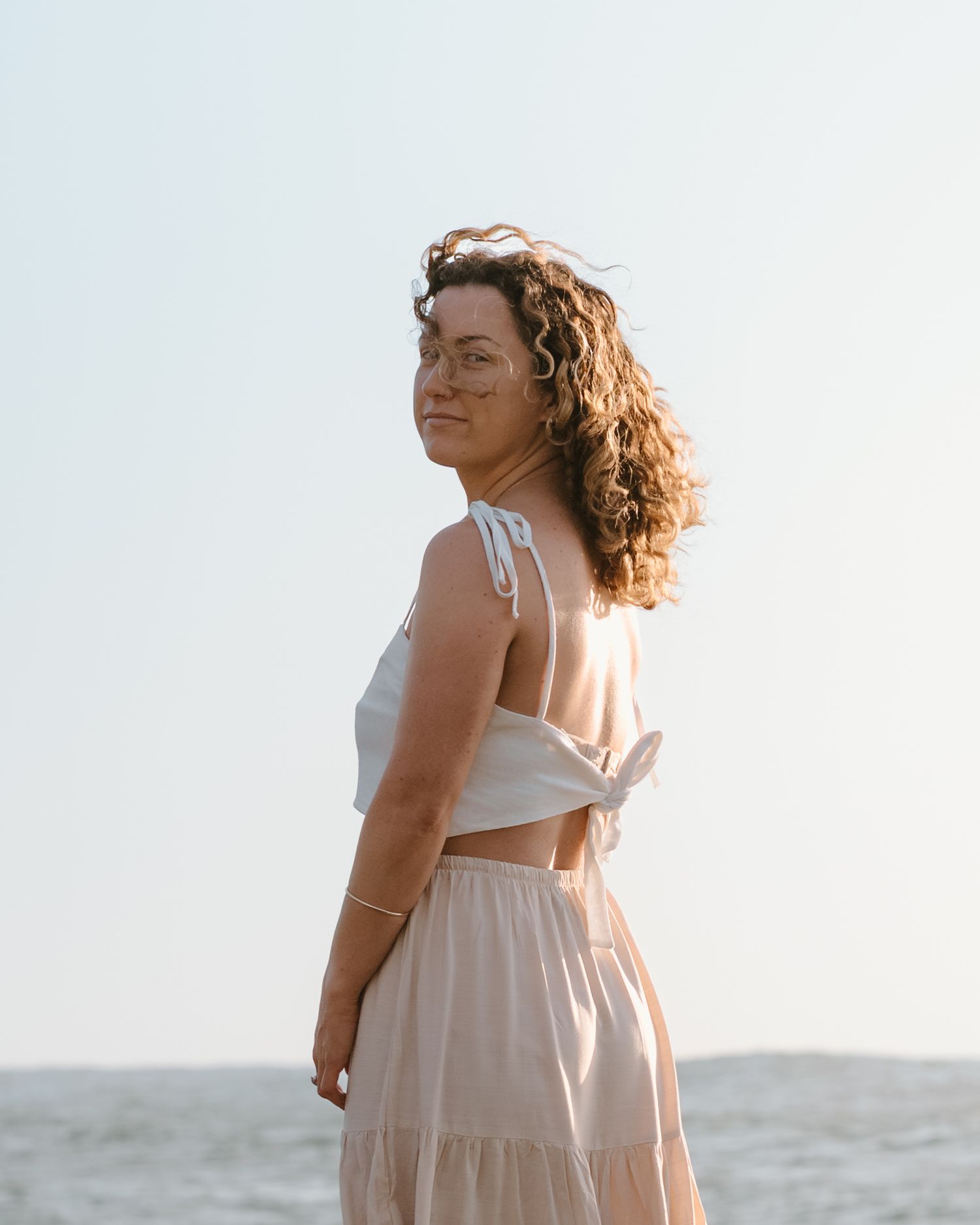 Woman in flowing white dress poses gracefully against hazy beach backdrop during golden hour sunset.