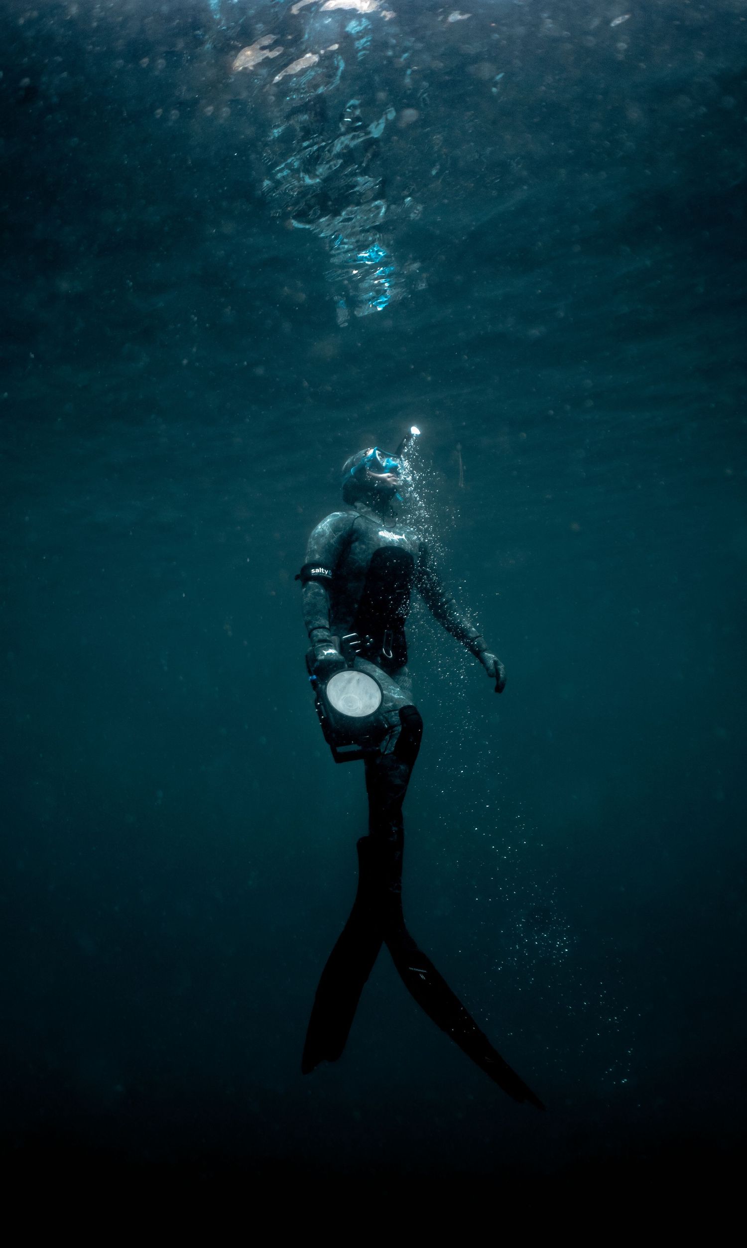 A scuba diver in a black wetsuit ascends through dark ocean water, holding a light, with bubbles rising around them.