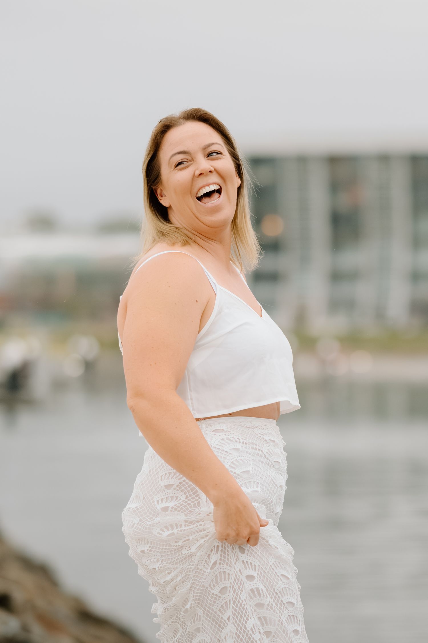 A joyful person in a white outfit laughs while posing near a waterfront on an overcast day.