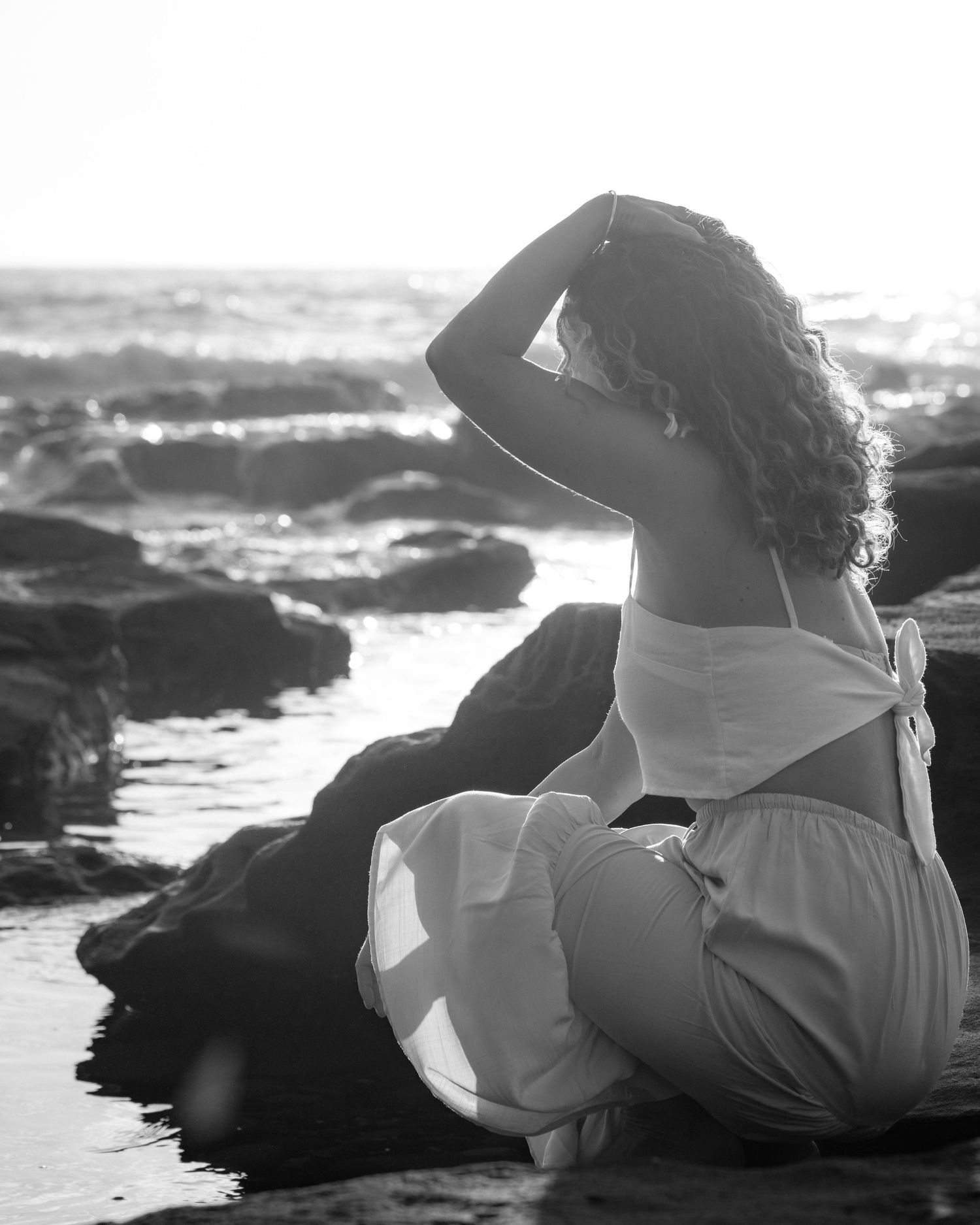 Black and white artistic photo series of a person in white dress reclining on coastal rocks by the ocean waves.