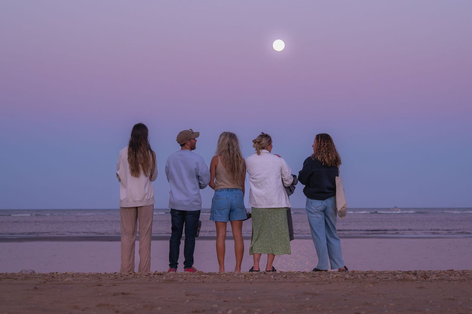 A group of friends sitting together on a beach at dusk watching the moon rise over the ocean horizon.