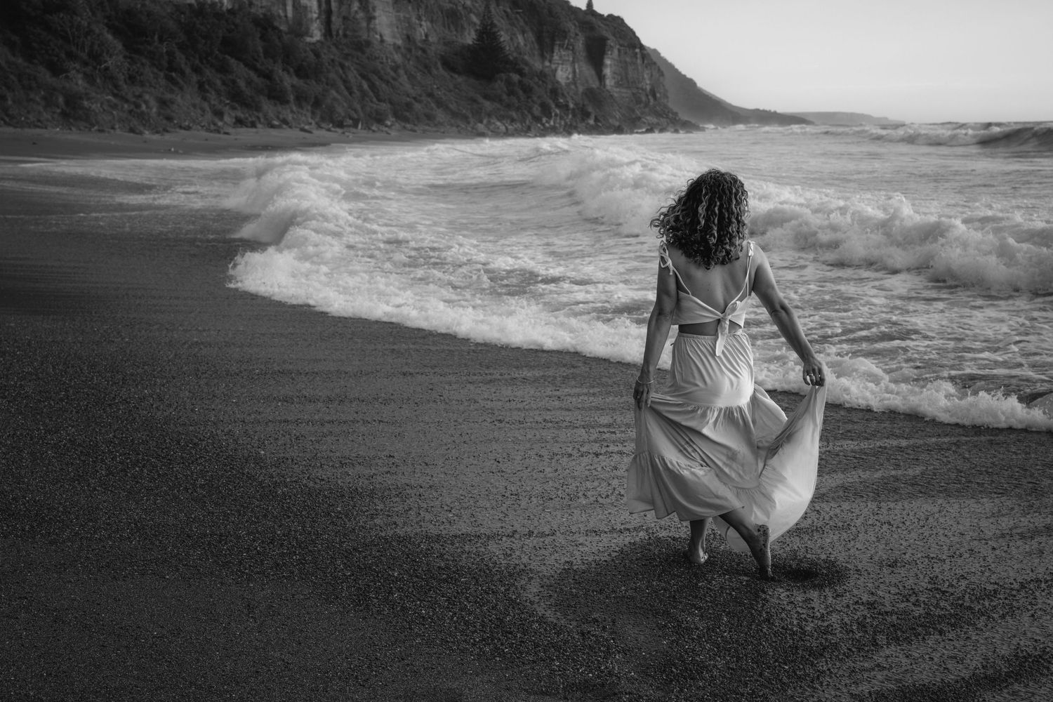 A black and white photo series showing someone in a flowing dress walking along a dramatic ocean shoreline.