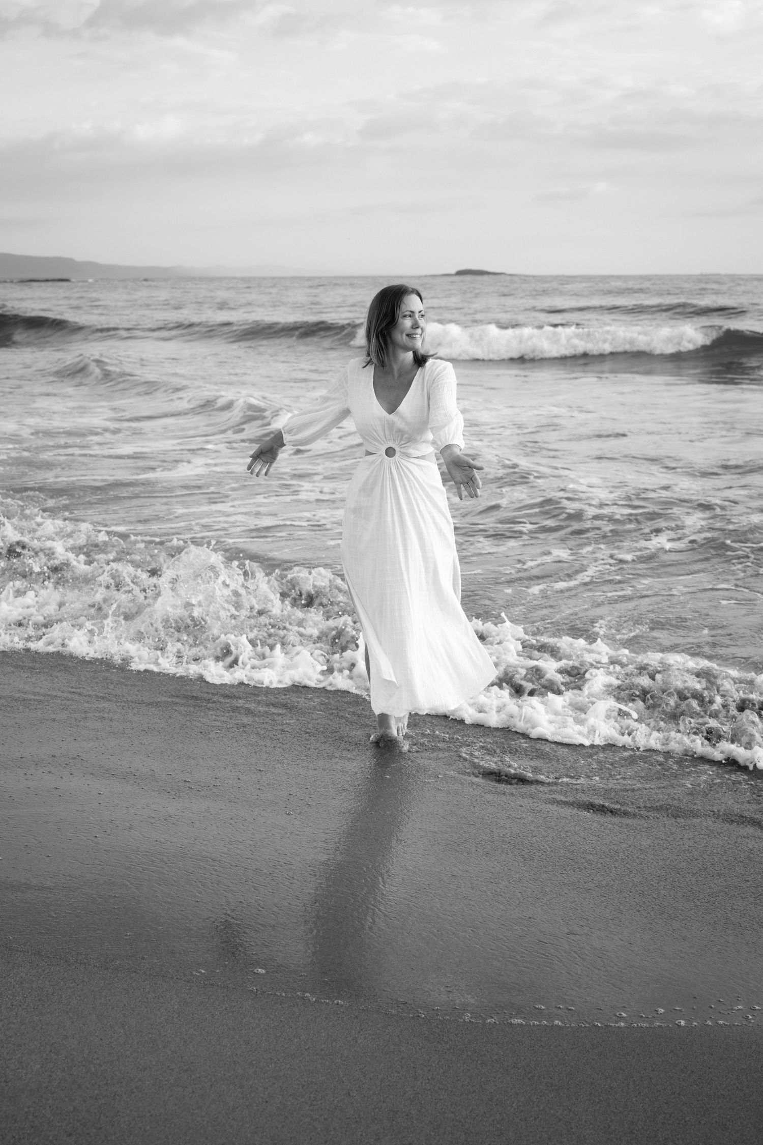 A person in a flowing white dress dances gracefully along a black sand beach as waves crash behind them.