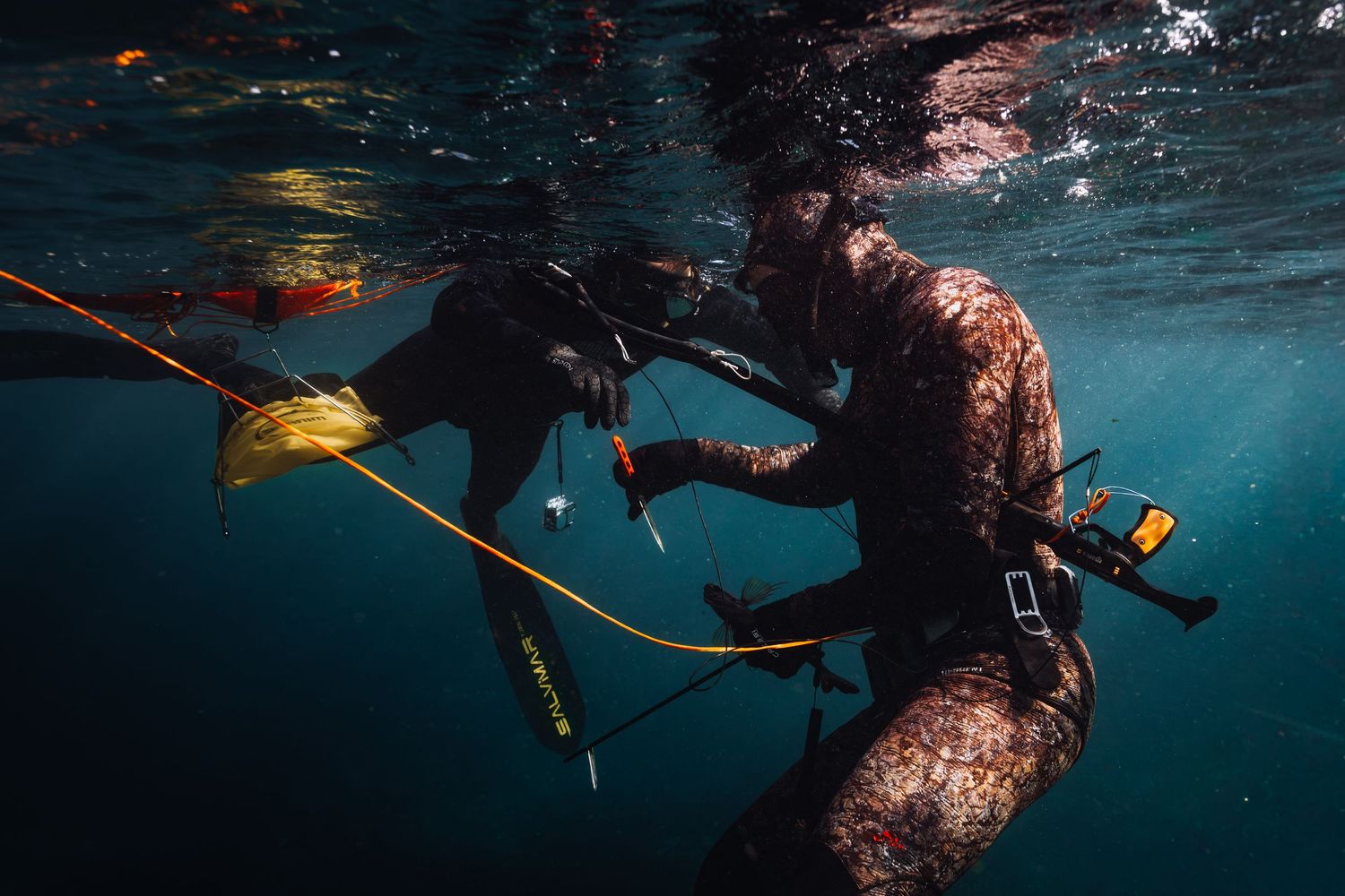Two divers in camouflage wetsuits spearfishing underwater, handling equipment with a yellow buoy line visible.