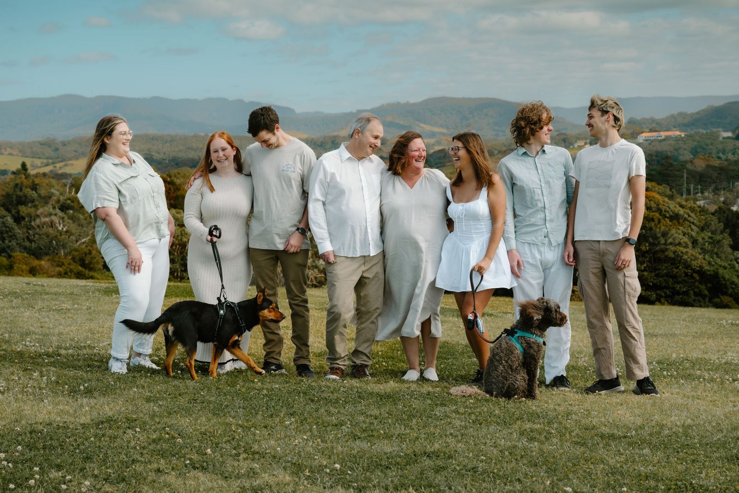 Family group with two dogs posing on green hillside with mountain views in white and neutral outfits.