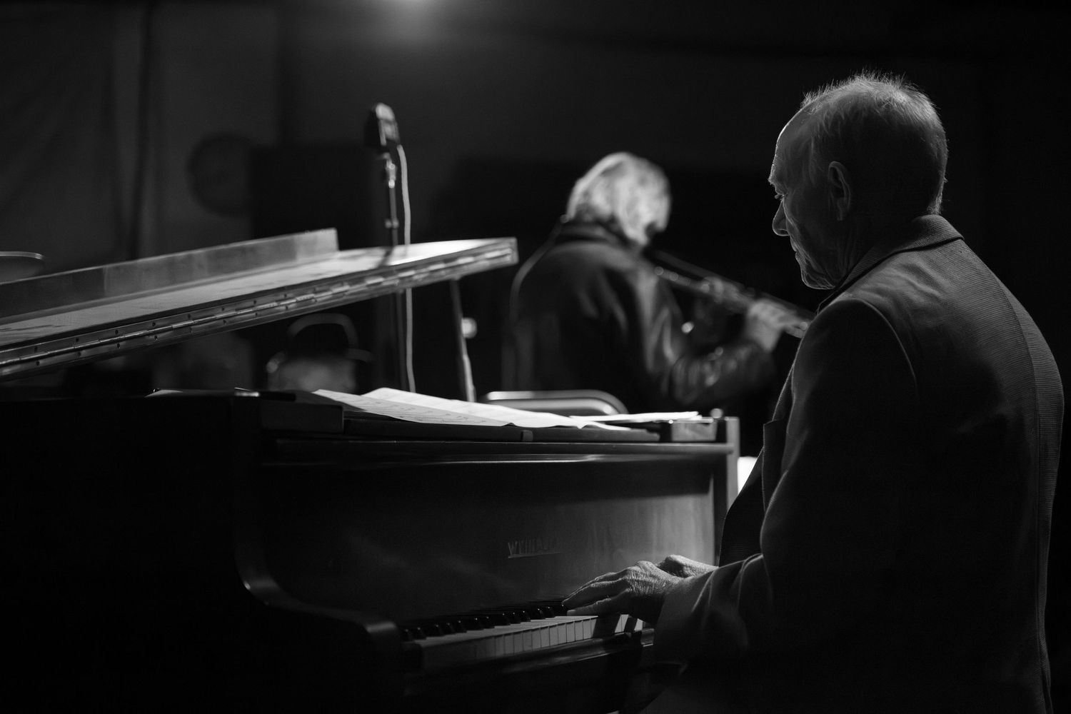 Black and white photo of an elderly man playing a grand piano on a dimly lit stage.