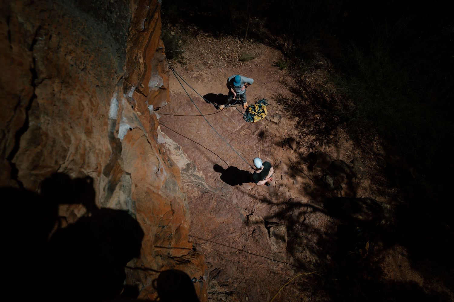 Aerial view of two rock climbers rappelling down a rugged red rock cliff face in a dark canyon.