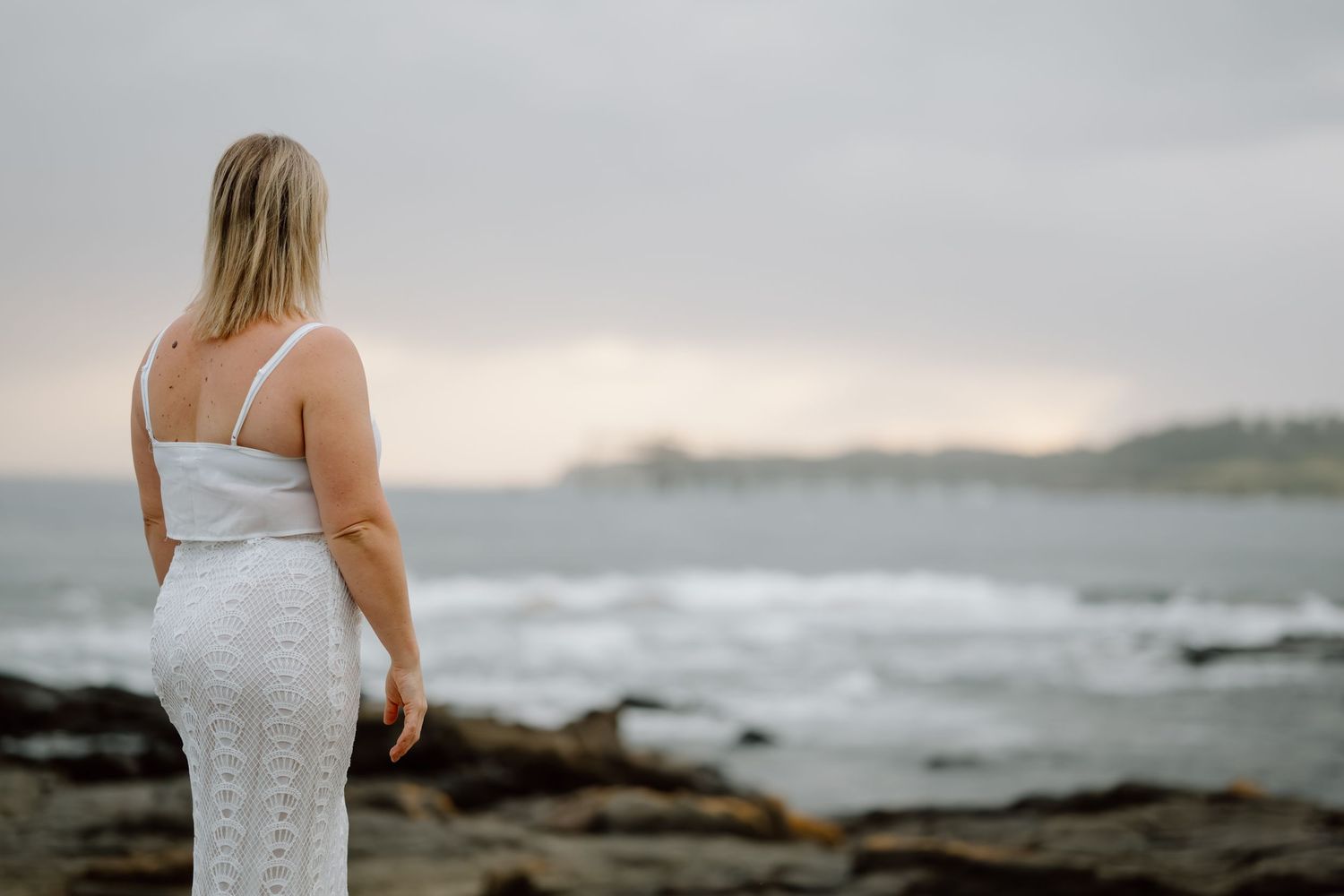 A woman in a white dress stands on a rocky beach at sunset with waves crashing in the background.