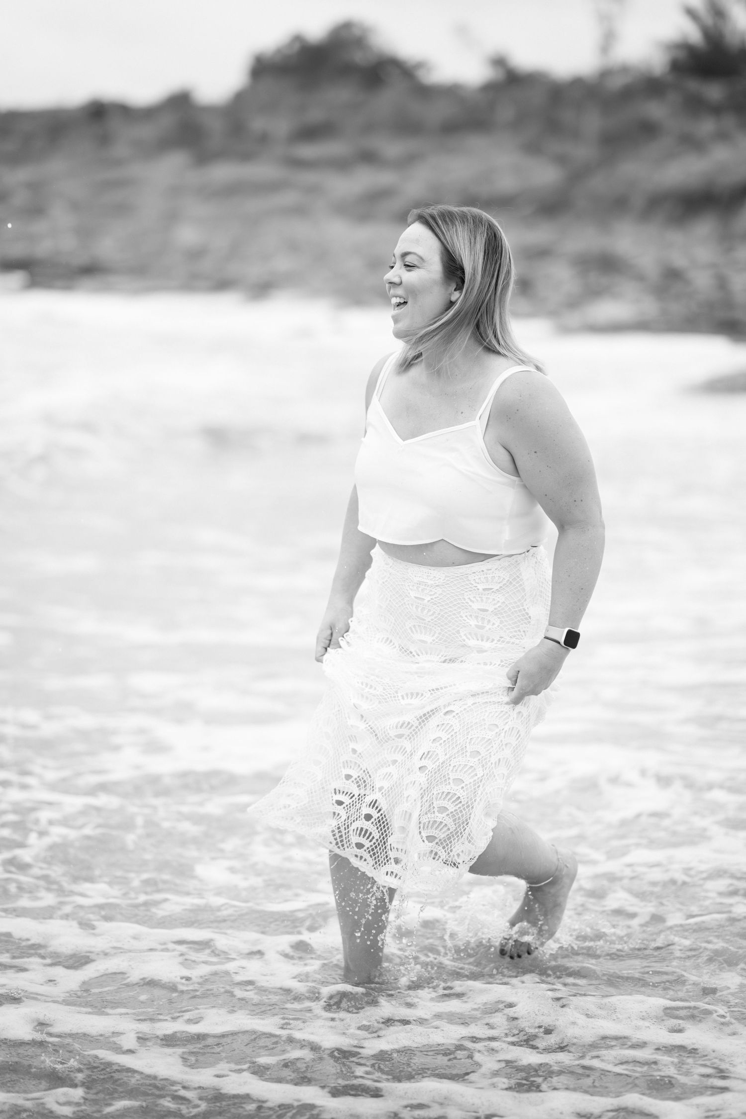 A person in a white dress splashes and plays in ocean waves at the beach in black and white photography.