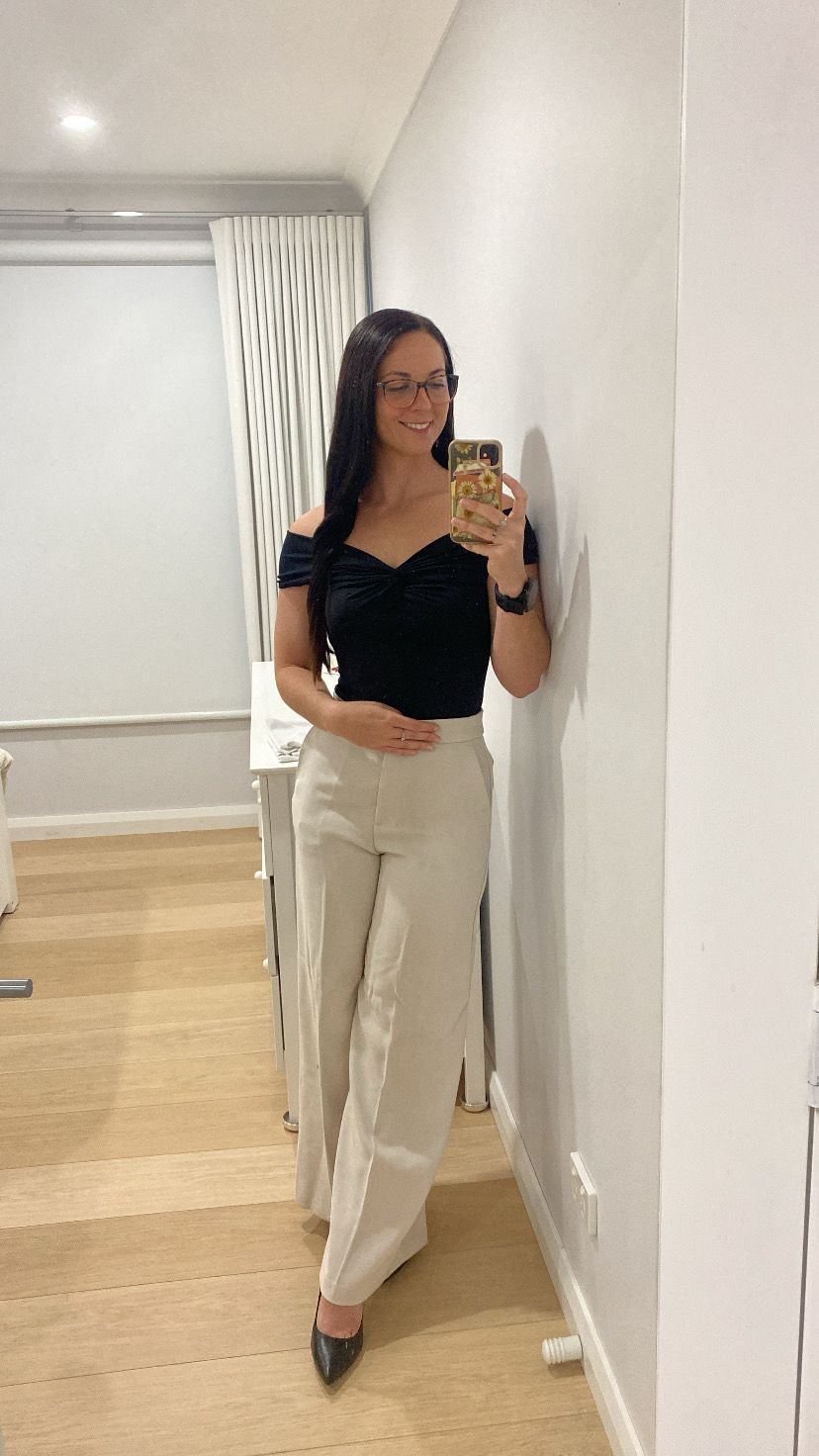 Woman in black off-shoulder top and beige wide-leg trousers takes mirror selfie in bright white room with wooden floors.