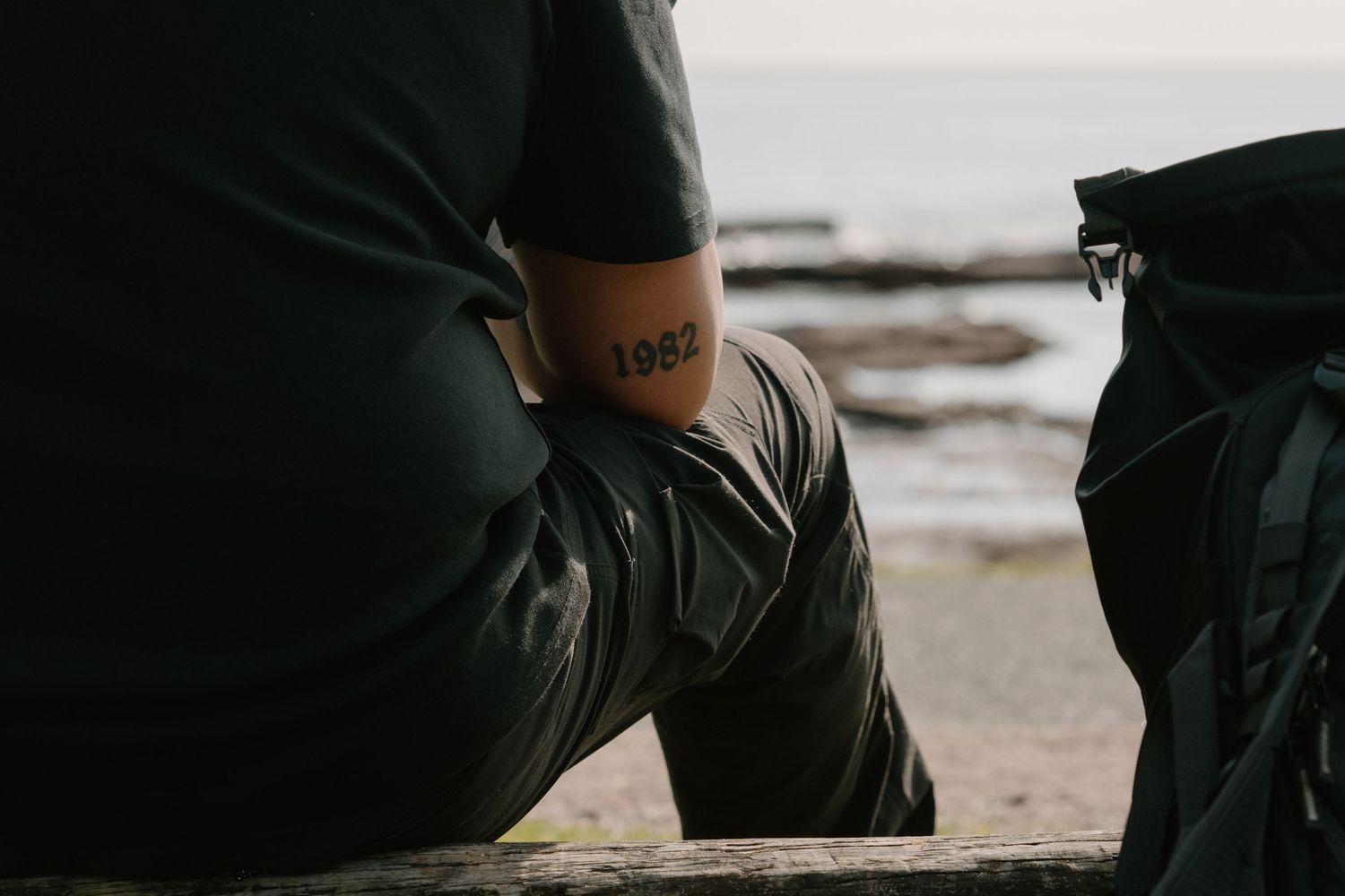 Person with 1982 tattoo on forearm sitting on bench near rocky beach with backpack beside them.