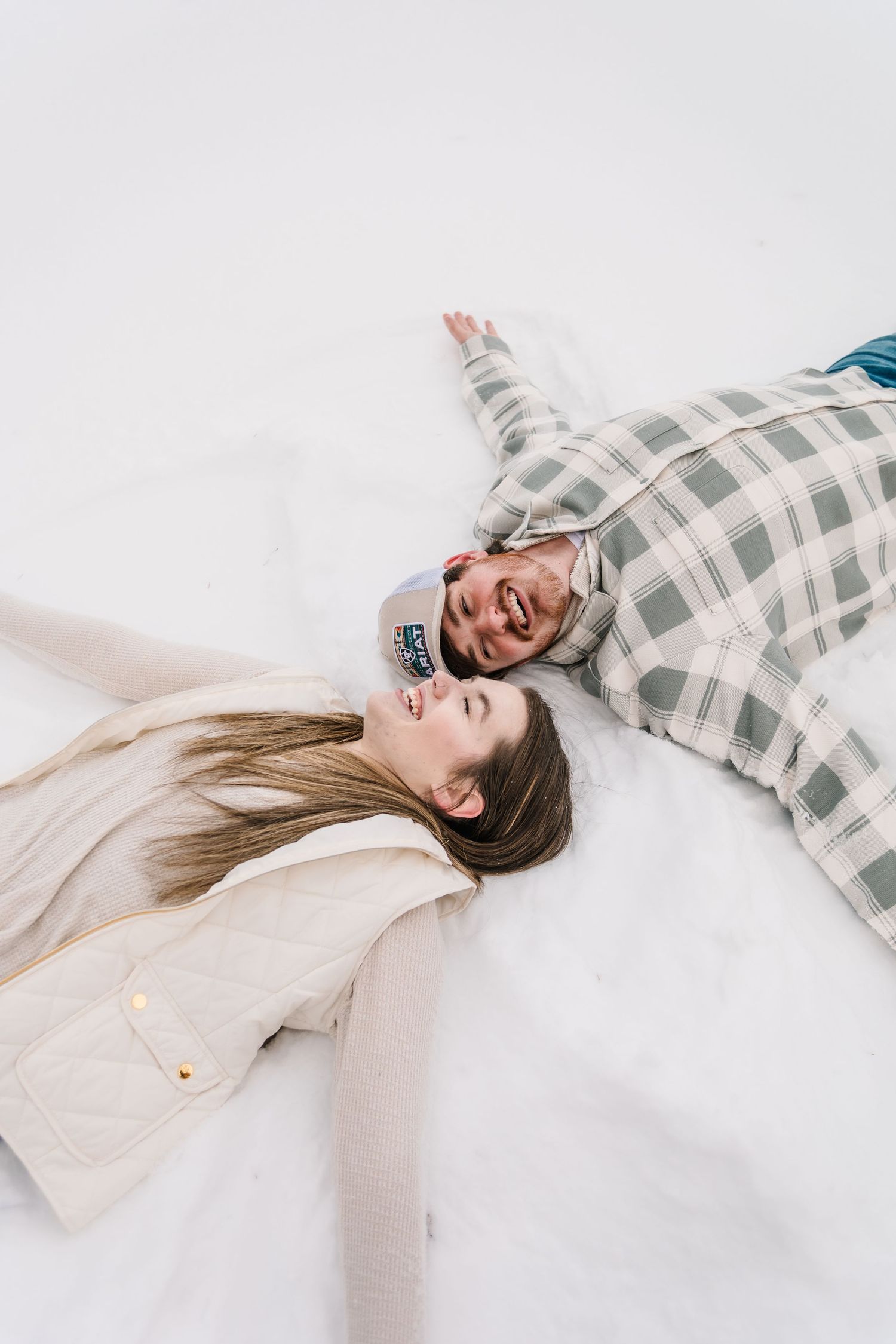 Couple laughing and making snow angels together while lying on white snow in winter outfits.