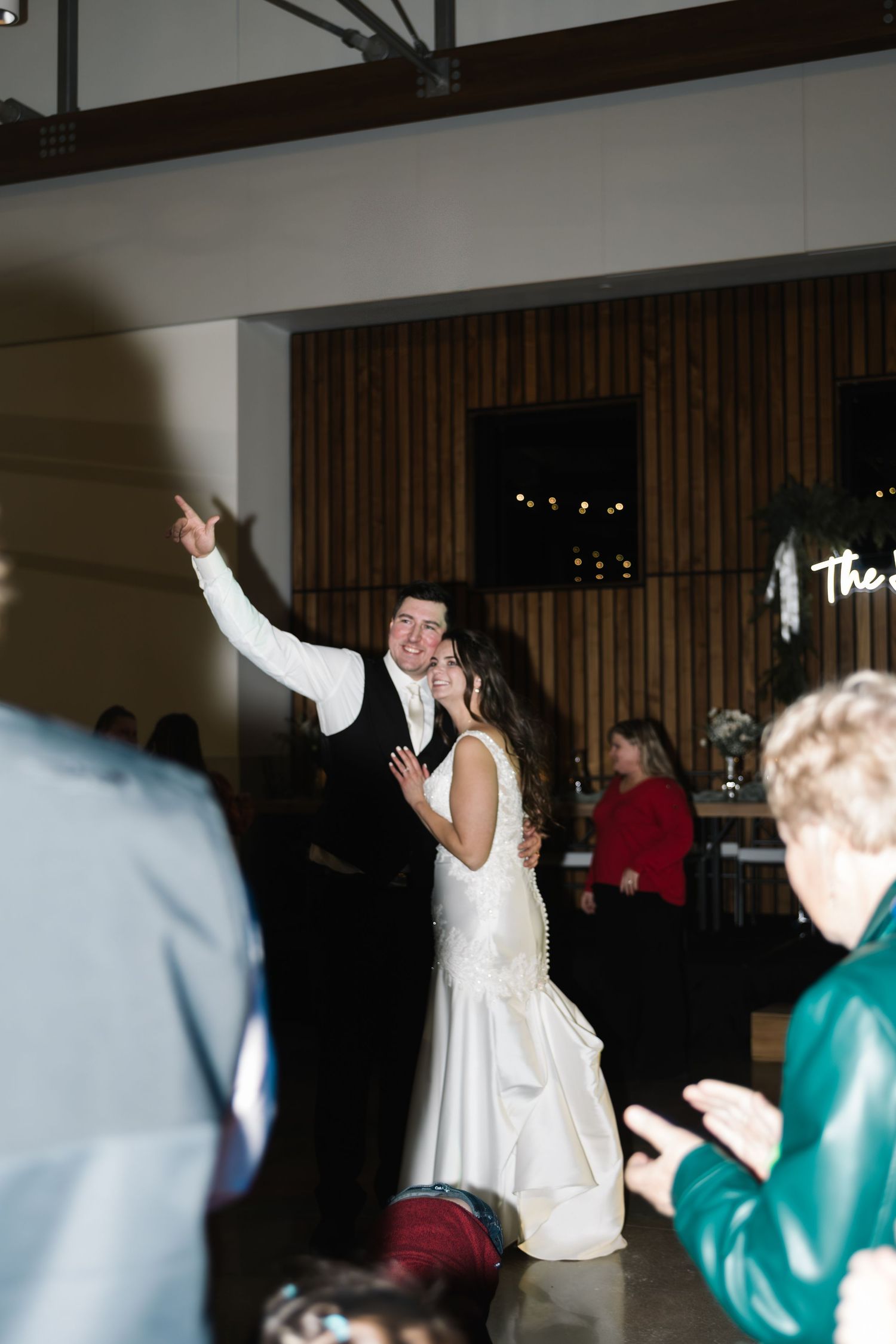 Bride and groom share first dance at wedding reception, groom pointing up joyfully while guests applaud around them.