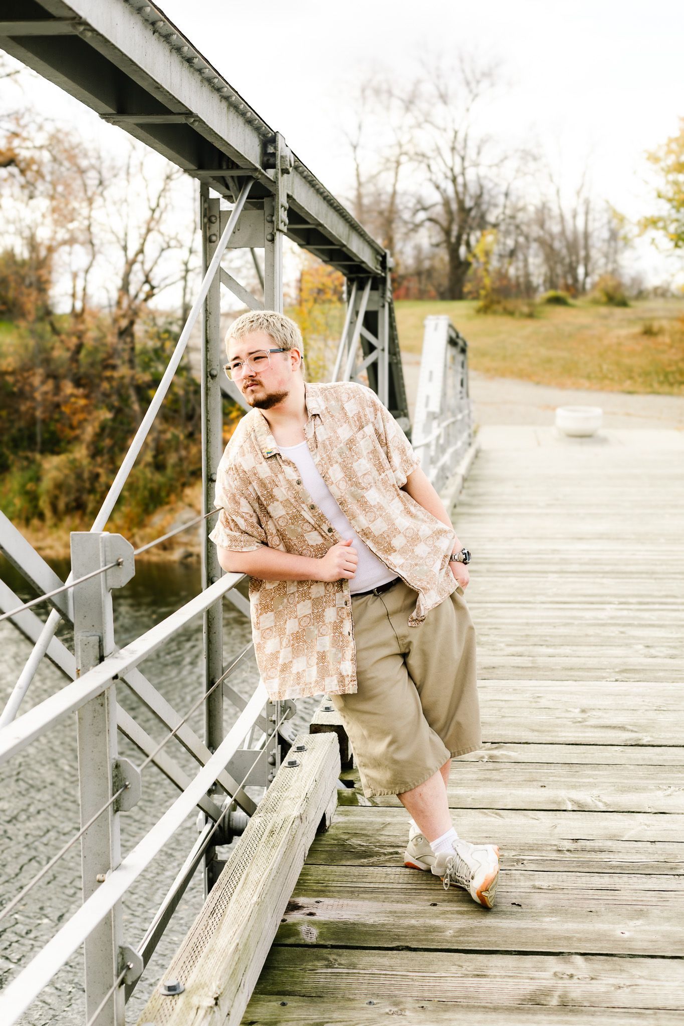 Young man in patterned shirt and khakis leaning on wooden bridge railing in autumn park setting.