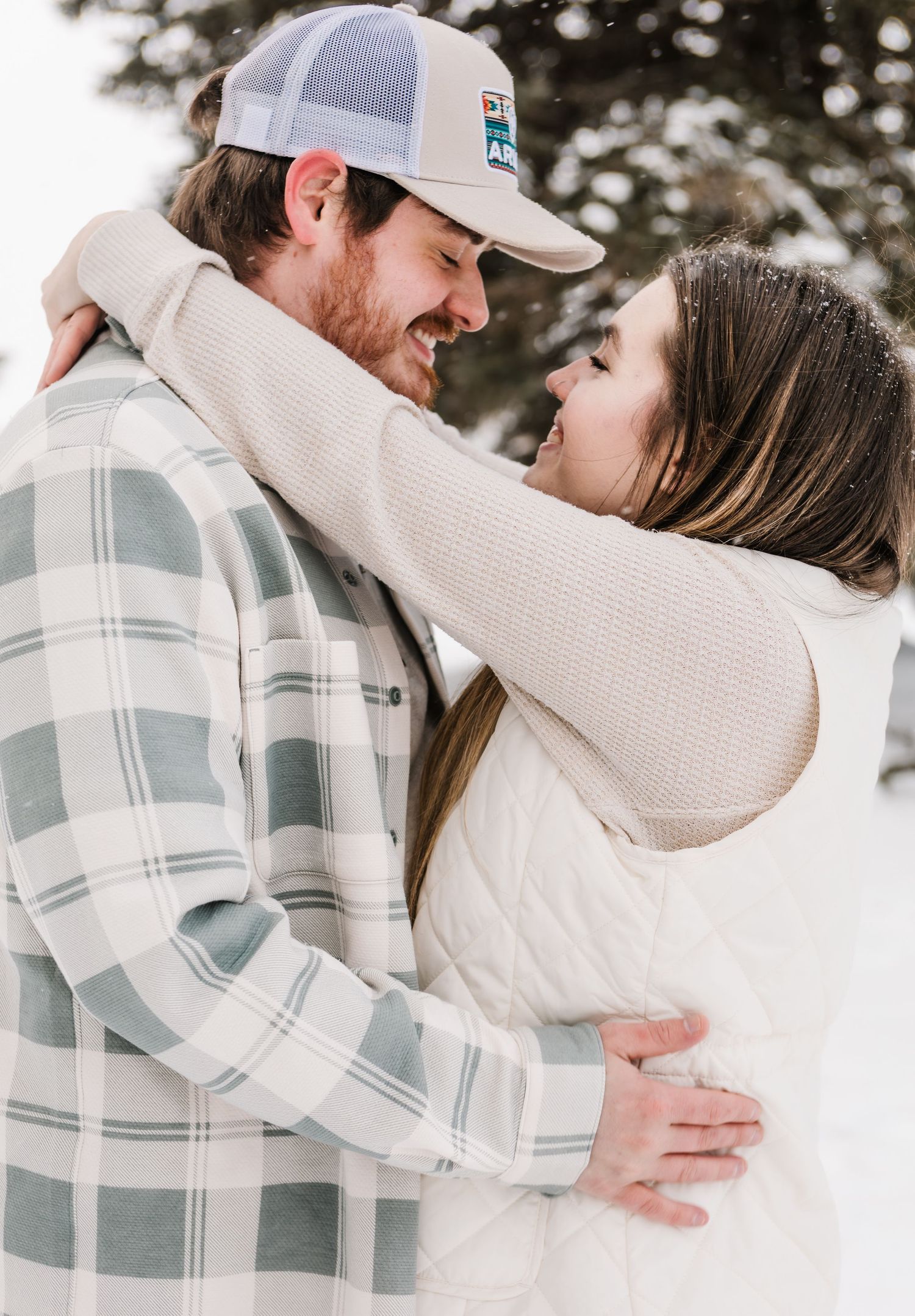 A couple embraces in a snowy winter setting, the man in a plaid shirt and cap, the woman in a white puffer jacket.