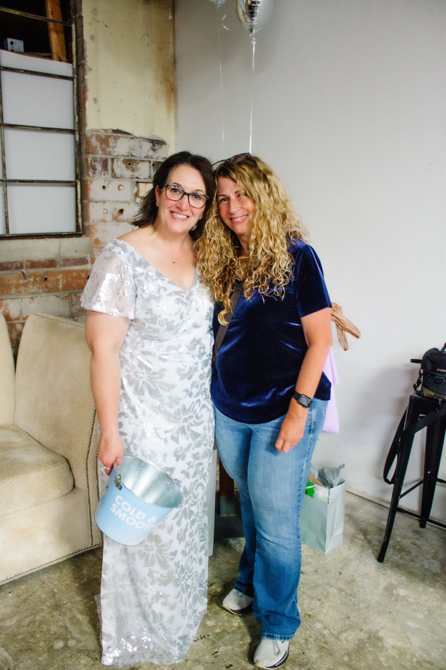 Two people posing together in a casual indoor setting with white walls and concrete floor.