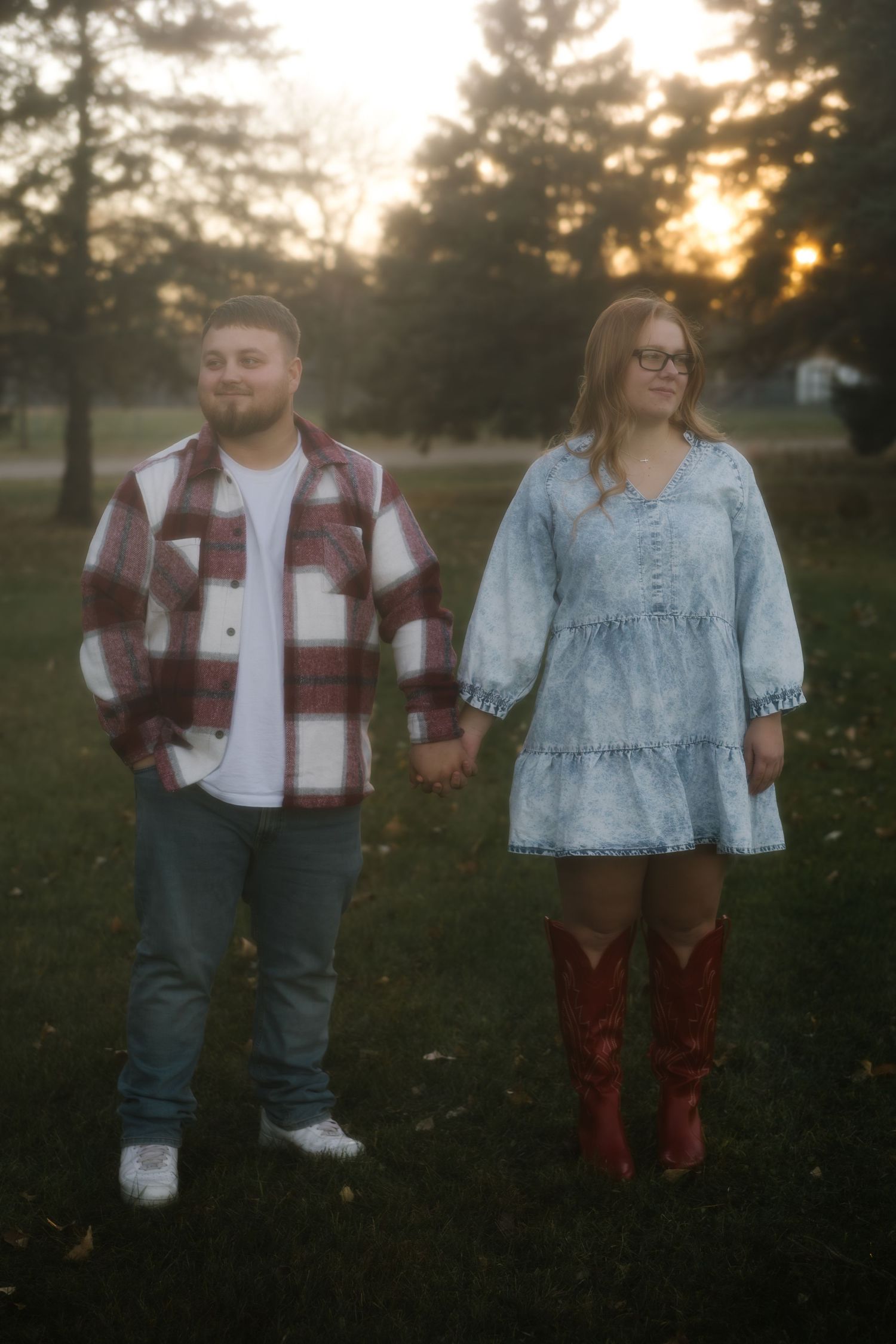 Couple holding hands at sunset in a park, man in plaid shirt and jeans, woman in denim dress and red cowboy boots.