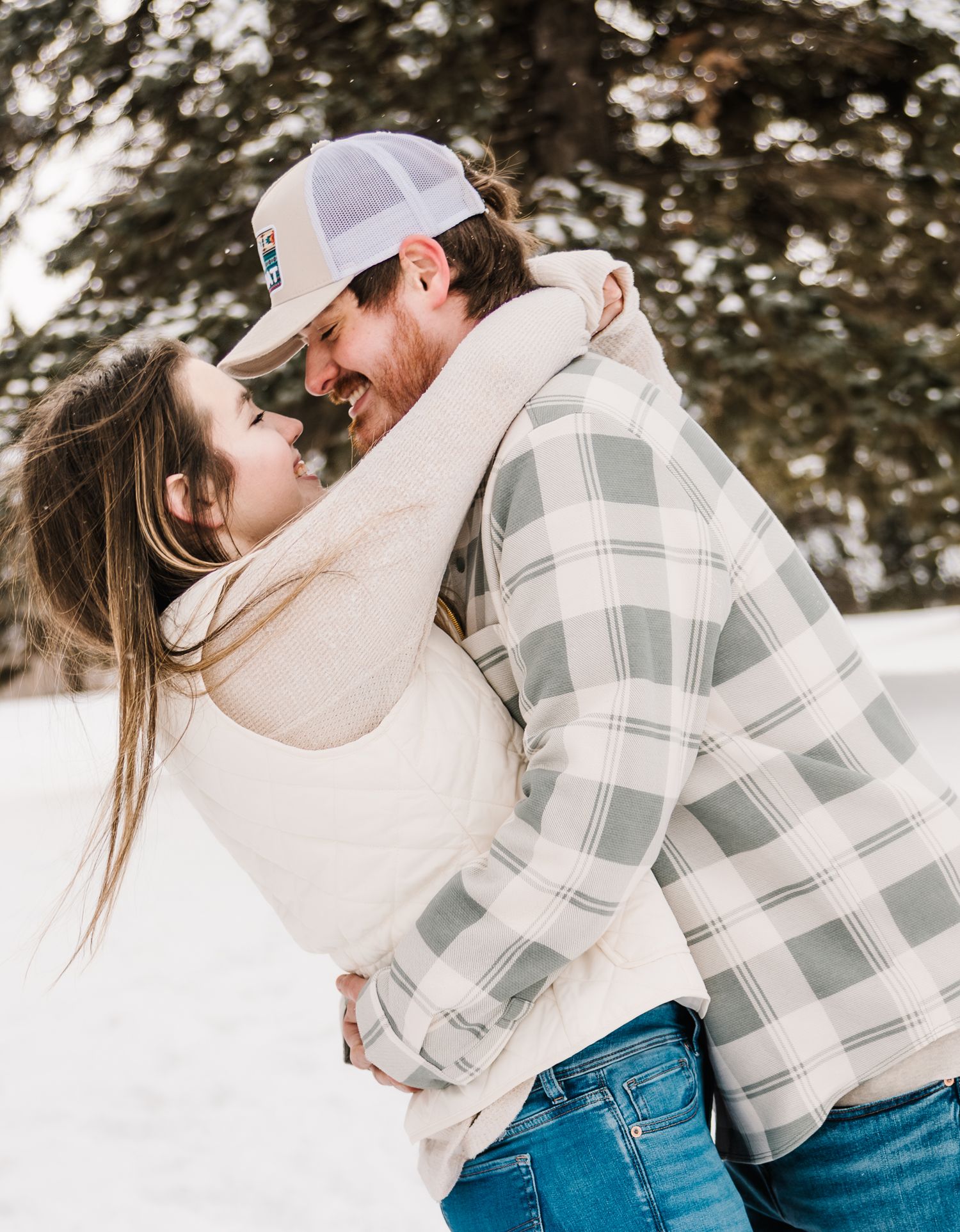 Couple embracing and smiling at each other in a snowy winter setting, wearing cozy plaid and cream sweaters.