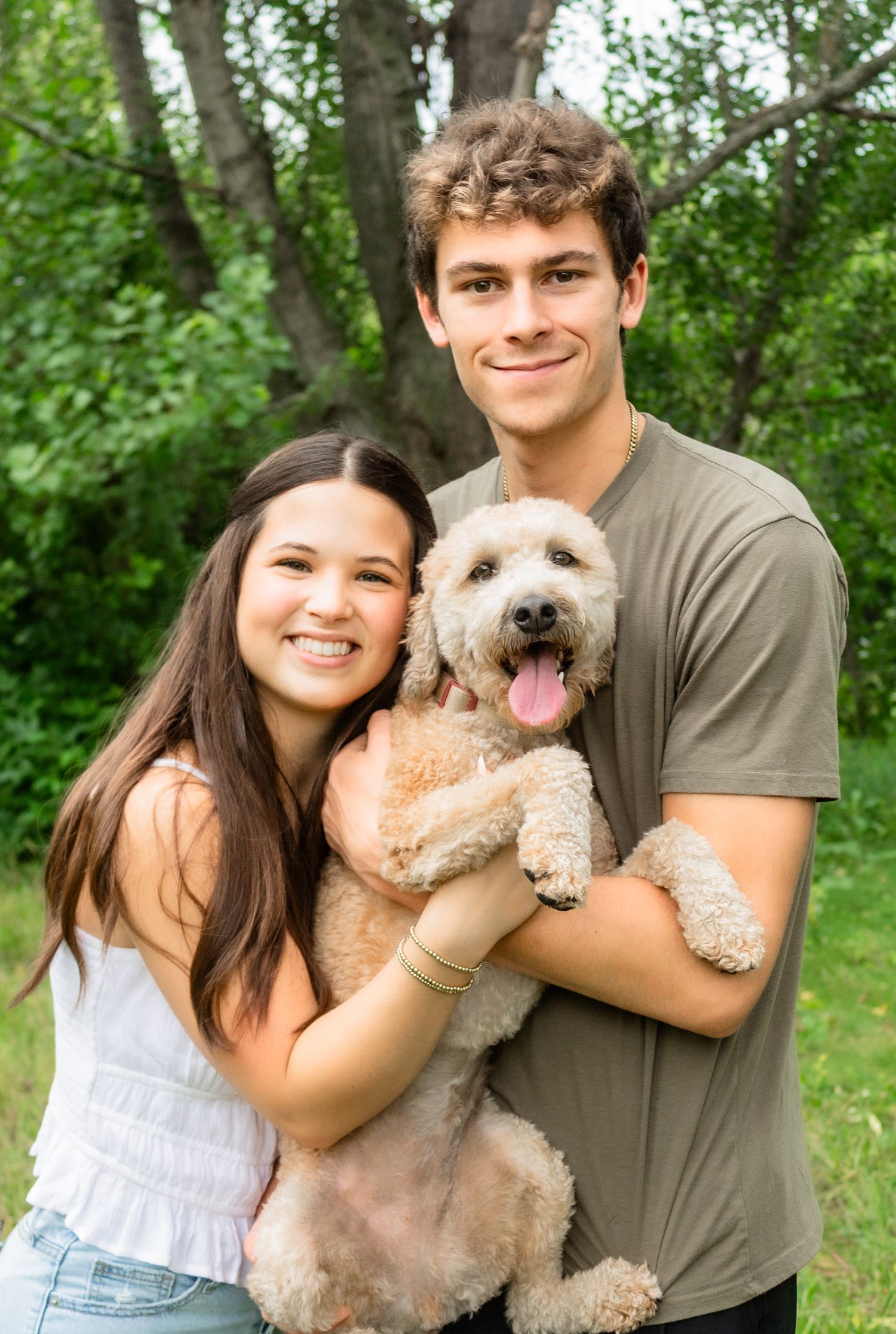 A happy couple holds their fluffy white dog while posing outdoors in a wooded area.