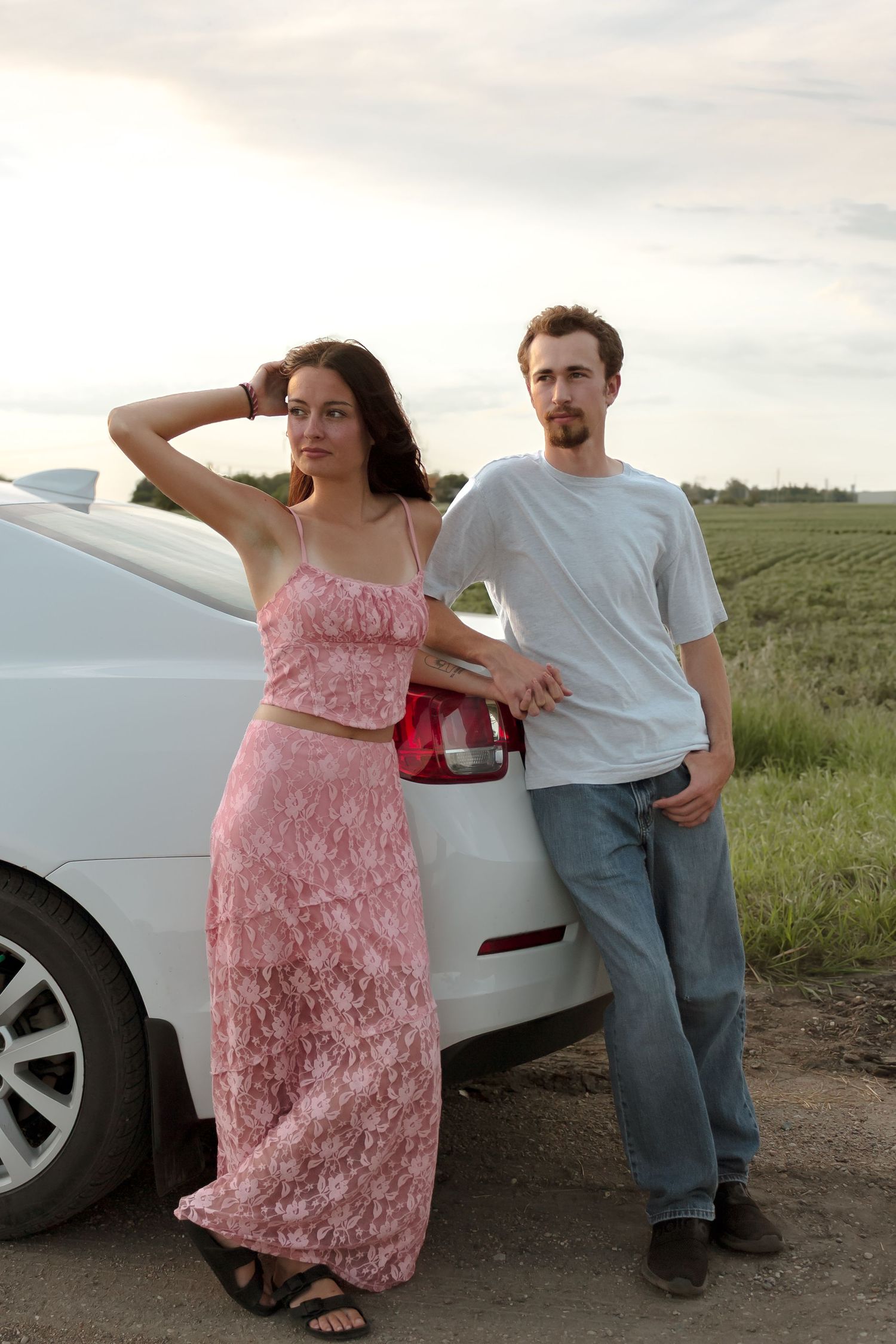 A couple poses casually against their white car in an outdoor setting.