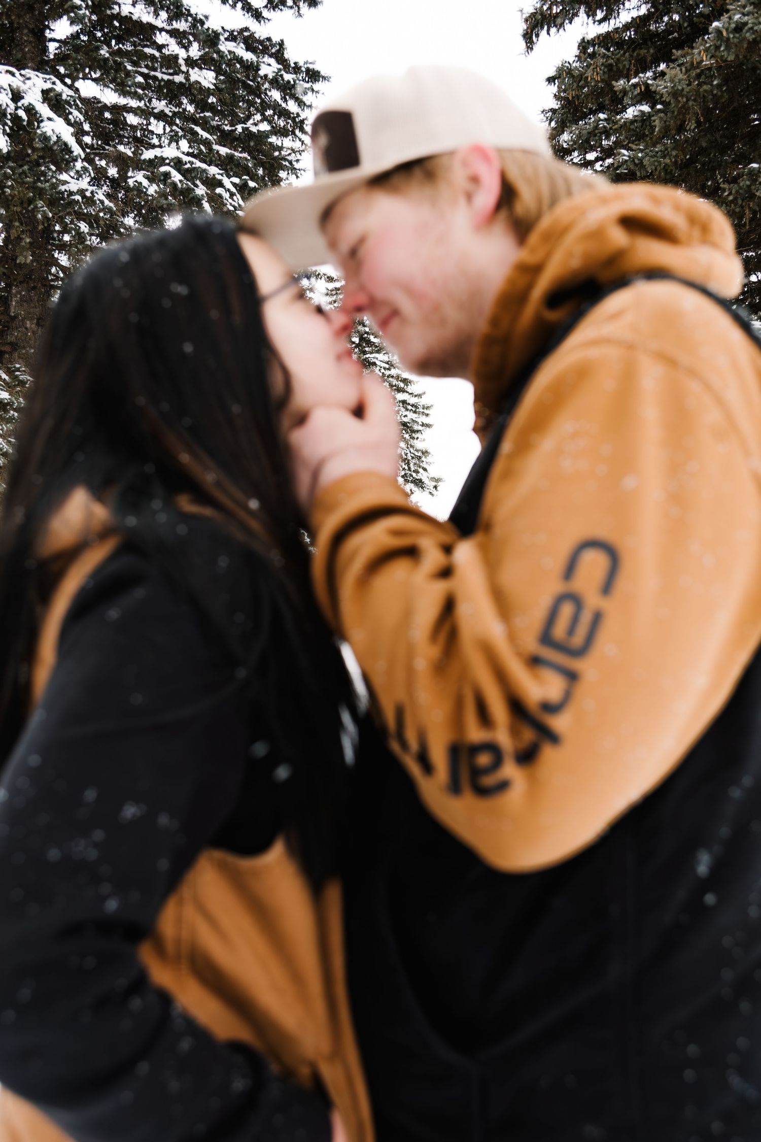 Couple kissing in snowy winter setting, man wearing Carhartt hoodie and cap, surrounded by snow-covered evergreen trees.