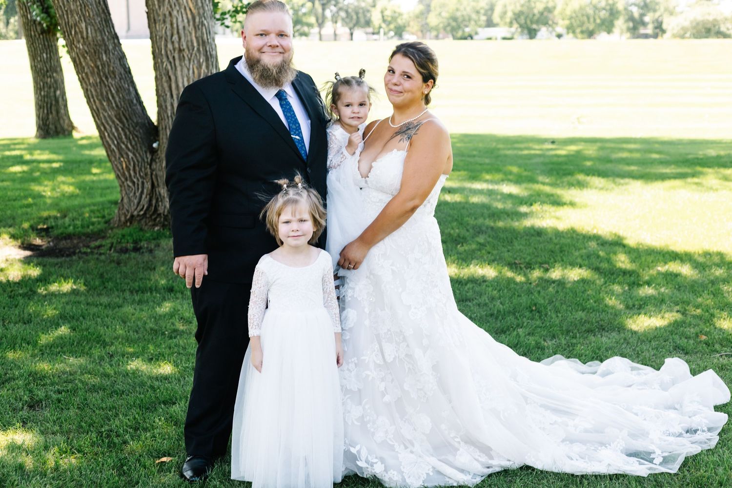 Bride in elegant lace gown poses with wedding party members on sunny lawn.
