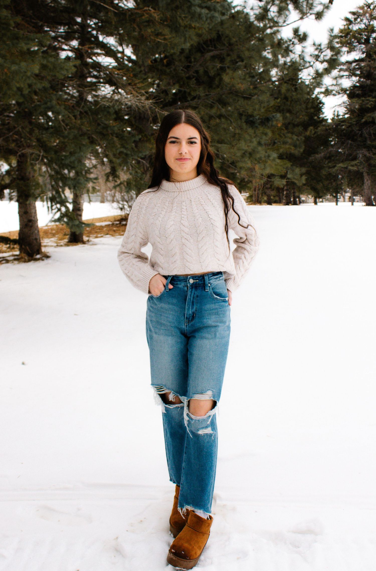 Casual winter outfit with brown boots, ripped jeans and knit sweater in snowy setting.