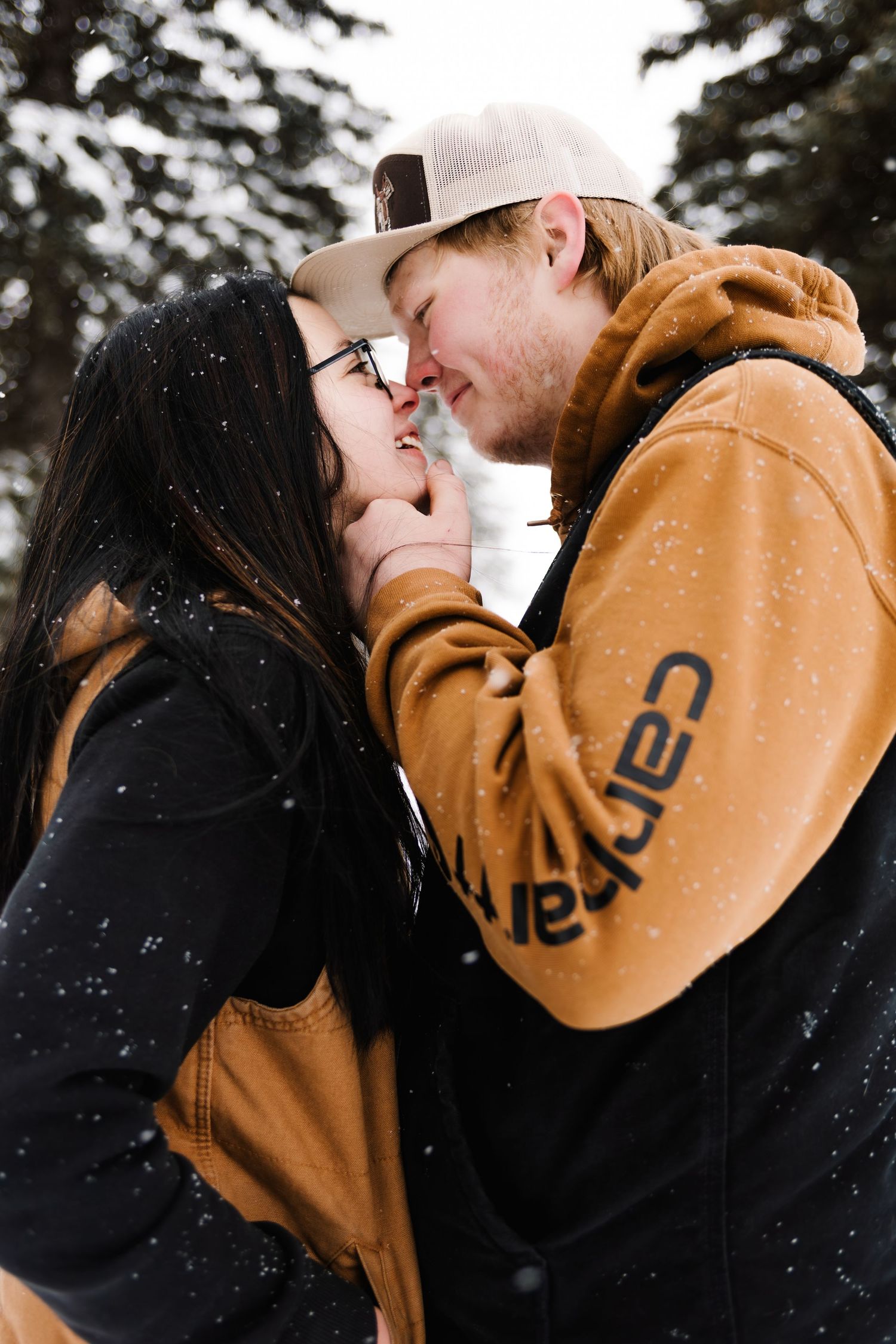 Couple sharing a romantic moment in the snow, wearing Carhartt jackets, with snowy pine trees in the background.